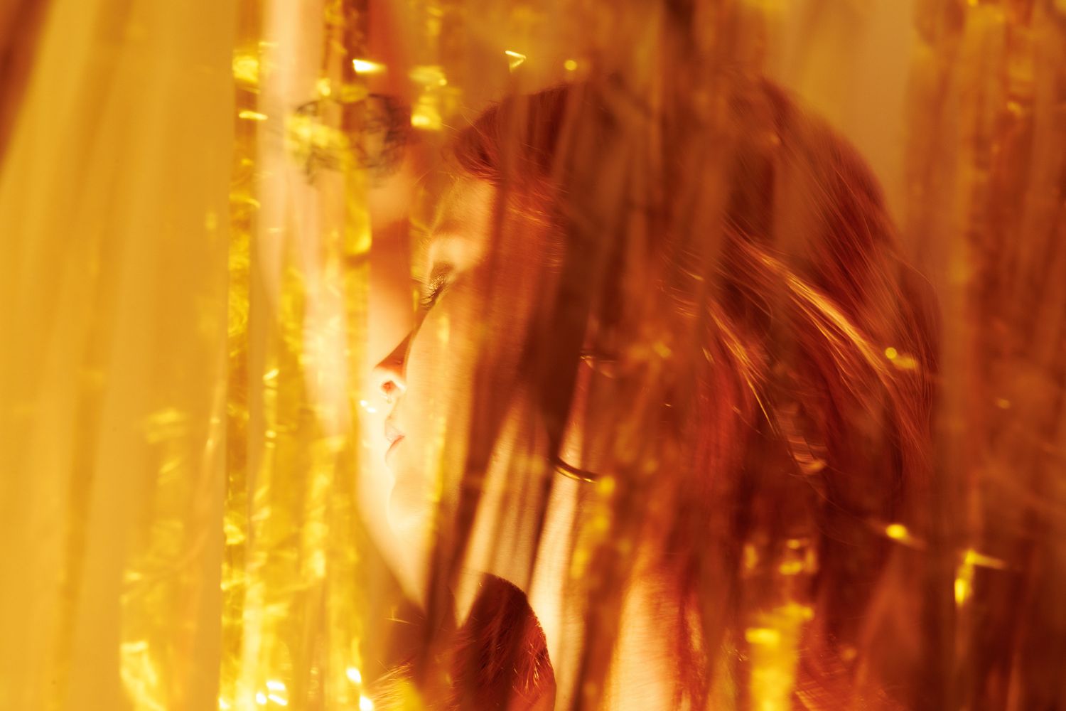 A woman partially obscured by golden tinsel curtains in warm amber tones.
