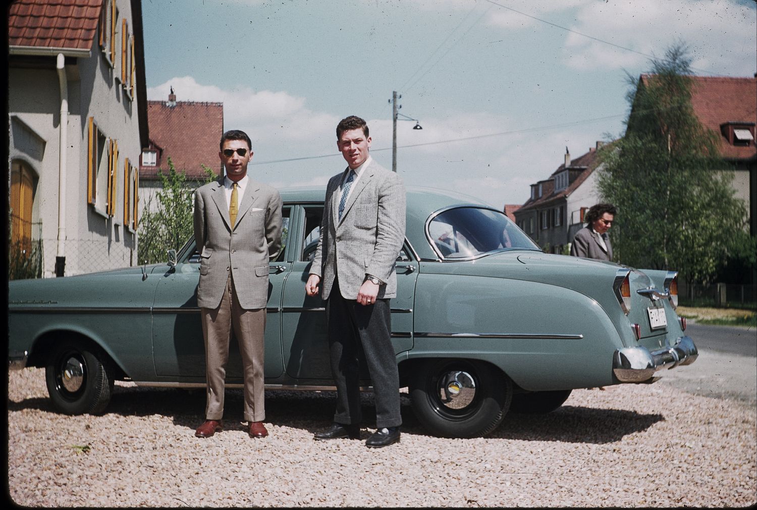 Two men in suits posing in front of vintage teal car on gravel driveway with buildings nearby.