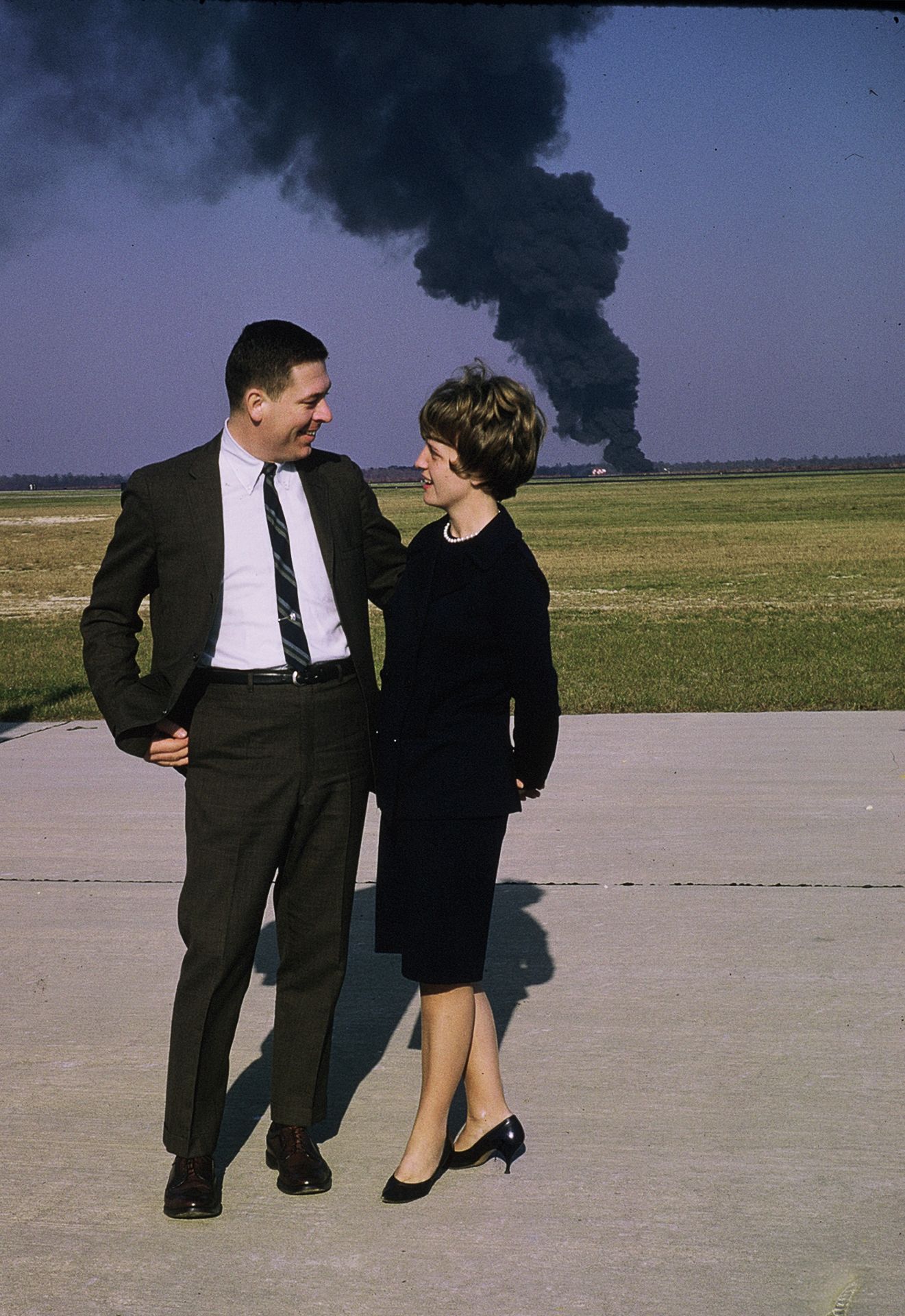 A man and woman in 1960s attire stand on a tarmac smiling, with a large black smoke plume rising in the background.