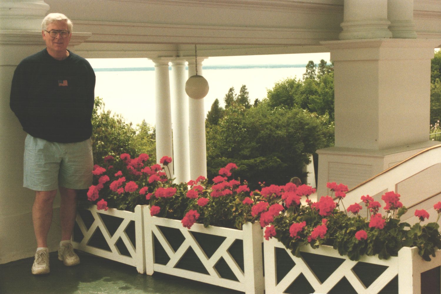 Man standing on white porch railing surrounded by vibrant pink geraniums at grand hotel.