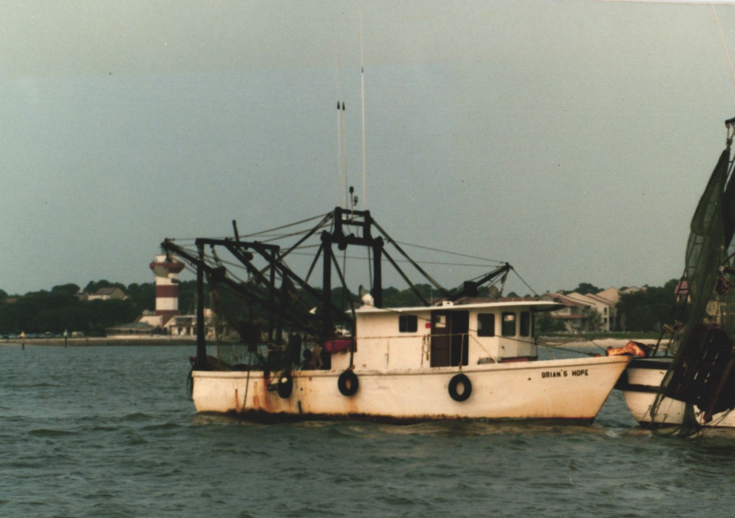A white commercial fishing boat with nets and rigging floats on calm harbor waters near a coastal town.