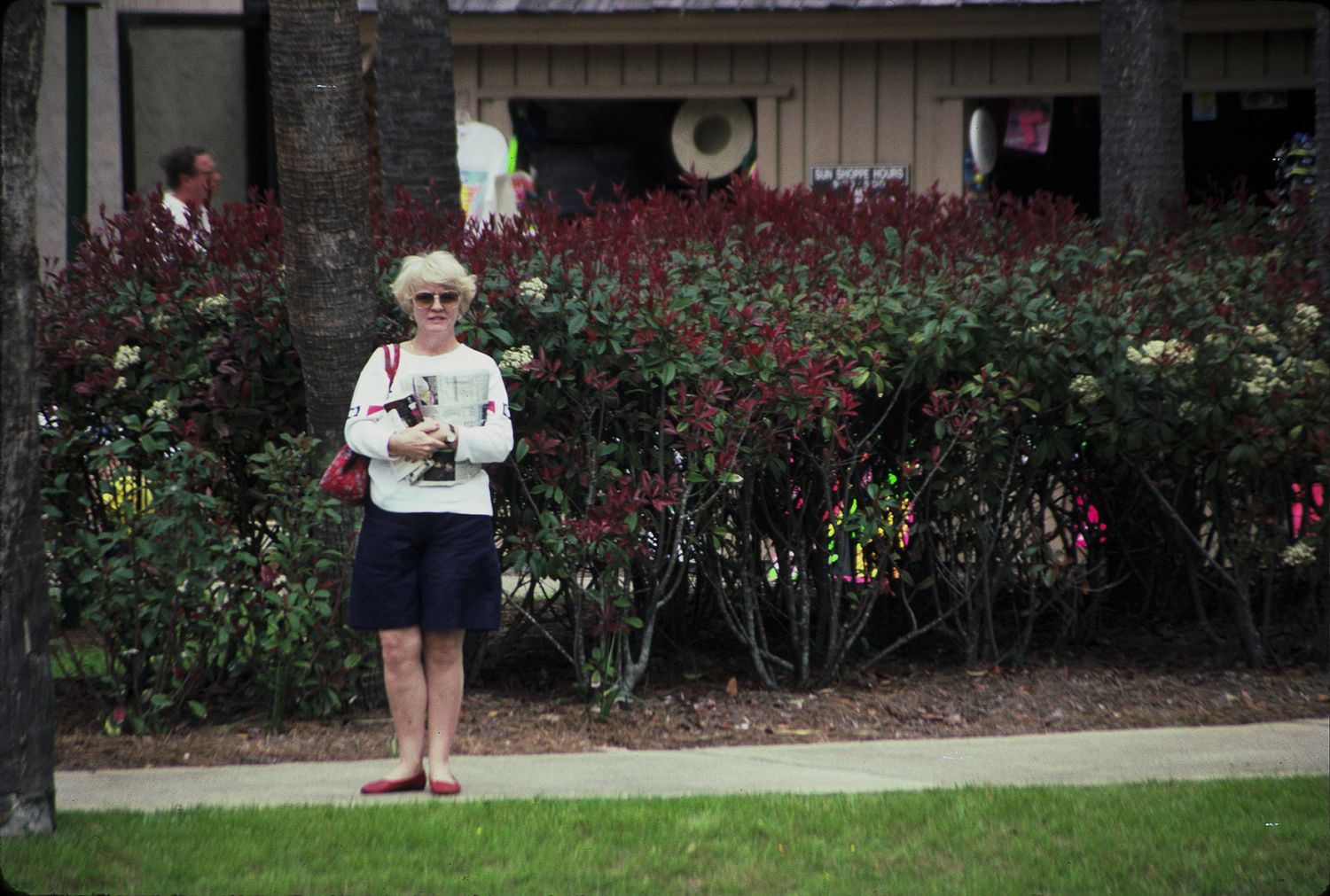 A woman in white top and blue shorts stands on a lawn in front of lush red flowering bushes near a house.