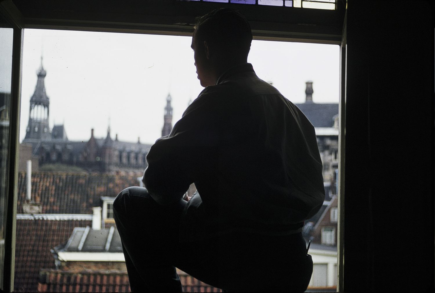A man sits silhouetted in a window overlooking European rooftops and a historic building.
