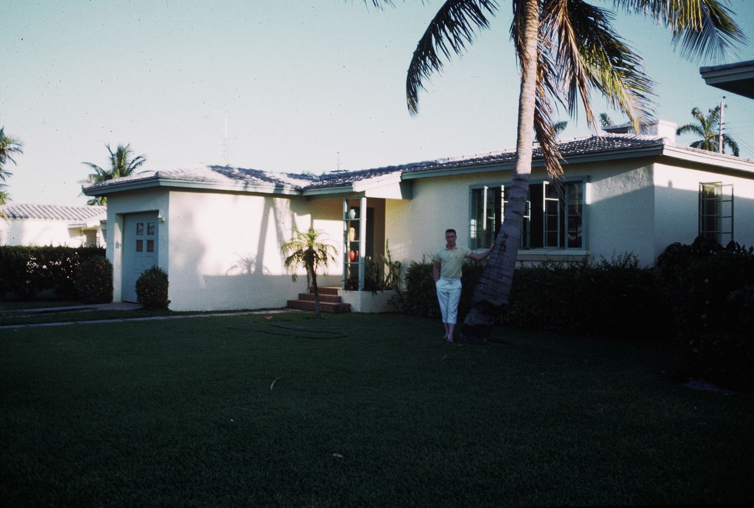 Person standing in front of a white mid-century ranch house with palm trees.