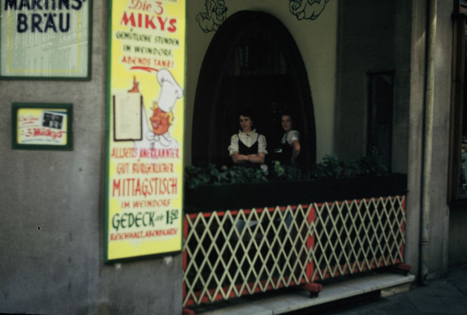 Two women in traditional German attire stand at a restaurant entrance with colorful signage for Martins Brau.