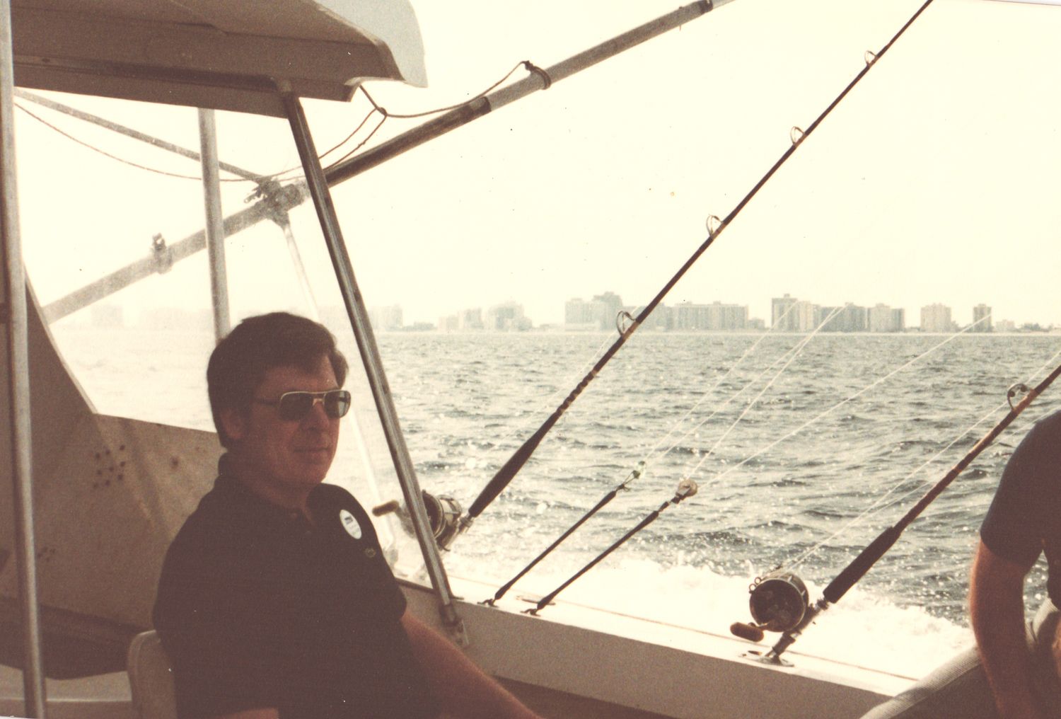 Man wearing sunglasses sitting on a fishing boat with rods and a city skyline visible across the water.