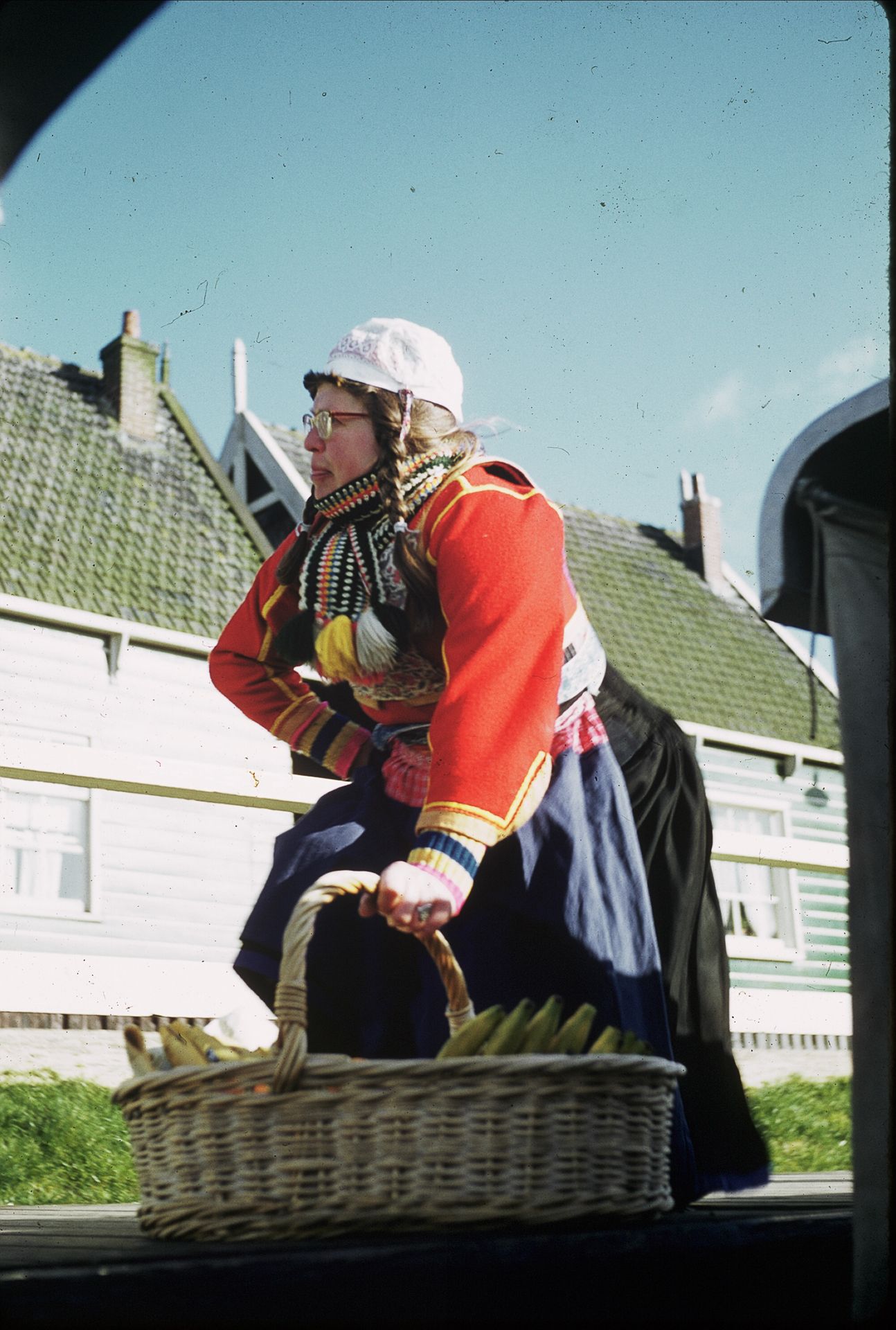 Woman in traditional Dutch costume carrying wicker basket near white wooden buildings under blue sky.