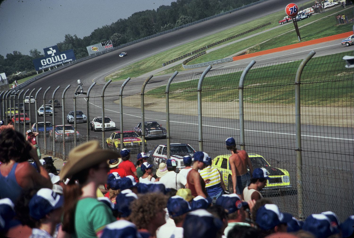 NASCAR race cars speed along a packed oval track while crowds of spectators watch from the grandstands on a summer day.