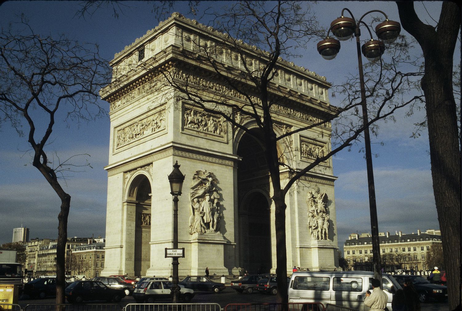The Arc de Triomphe in Paris surrounded by trees and traffic on a clear day.