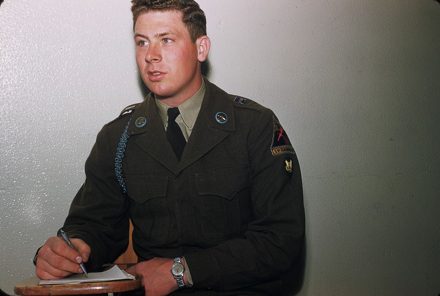 US Army soldier in olive green uniform with tie writes notes at a desk, wearing a wristwatch.