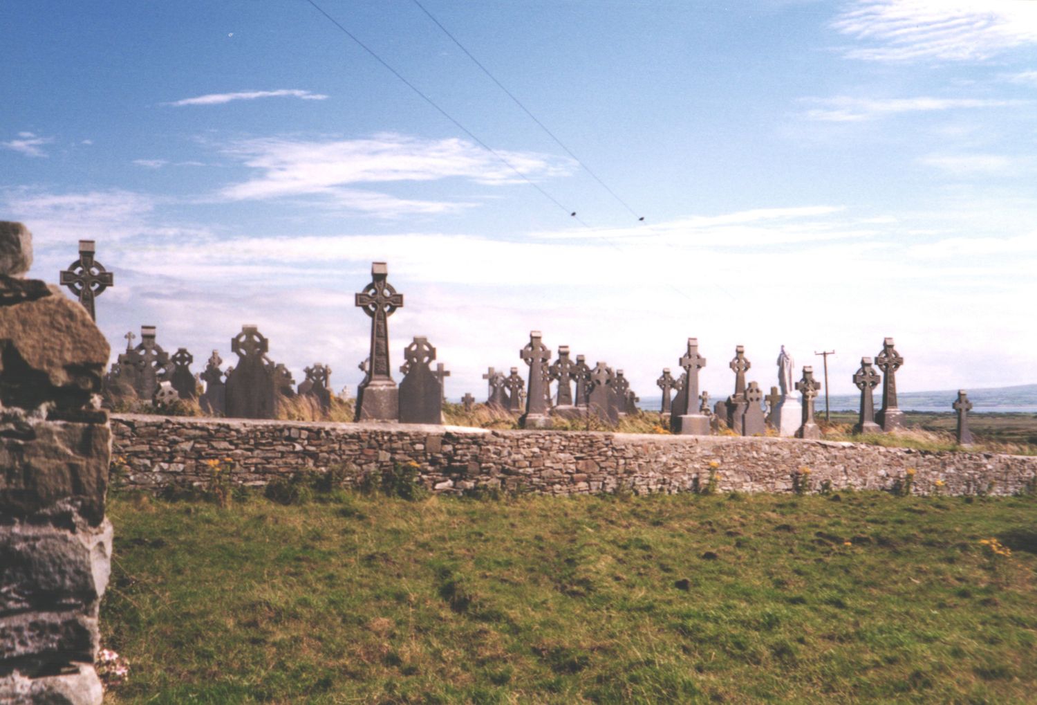 Ancient Celtic cross cemetery with stone wall enclosure under blue sky in rural Ireland.