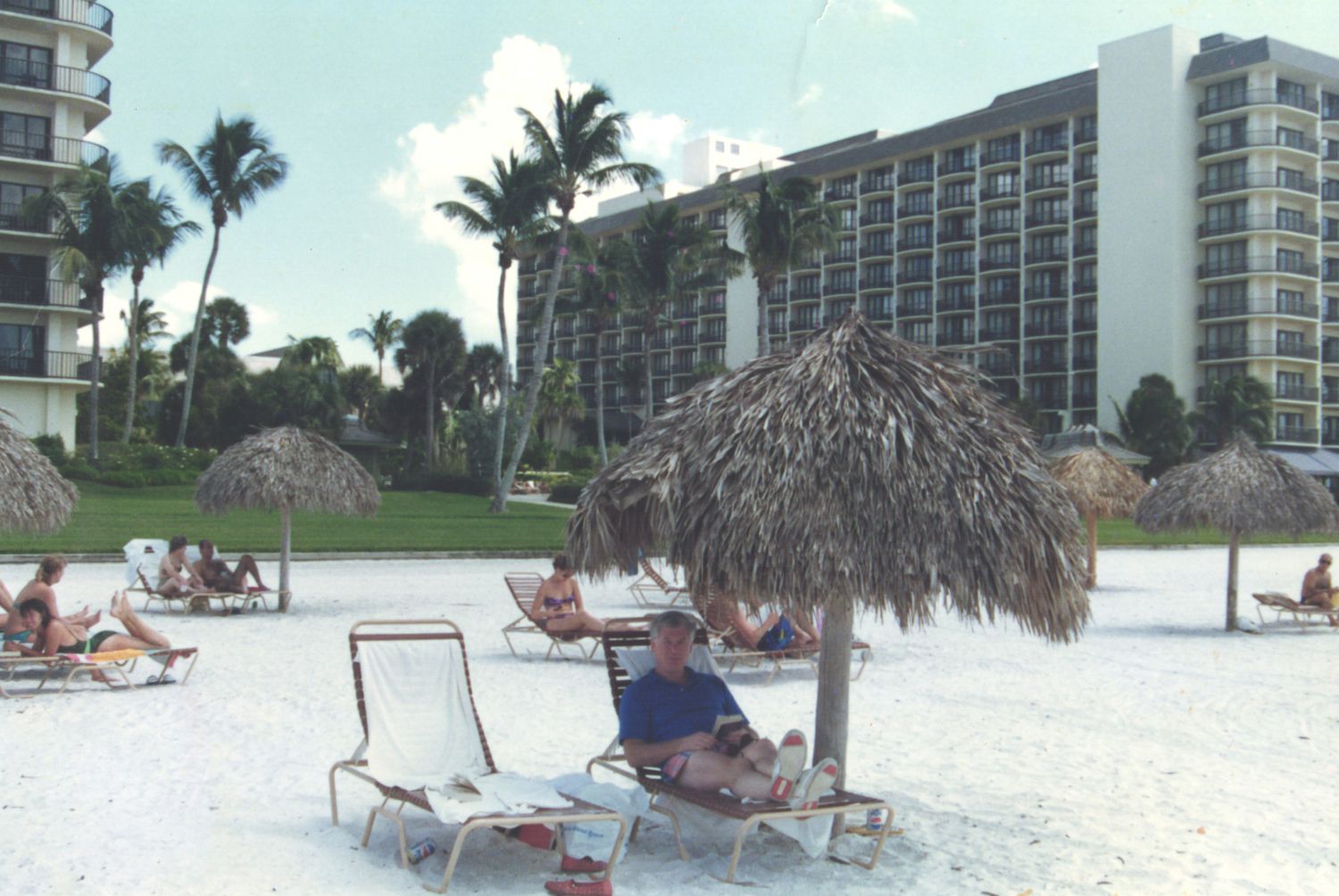 A man relaxes on a lounge chair under a thatched palapa umbrella on a white sandy beach resort.