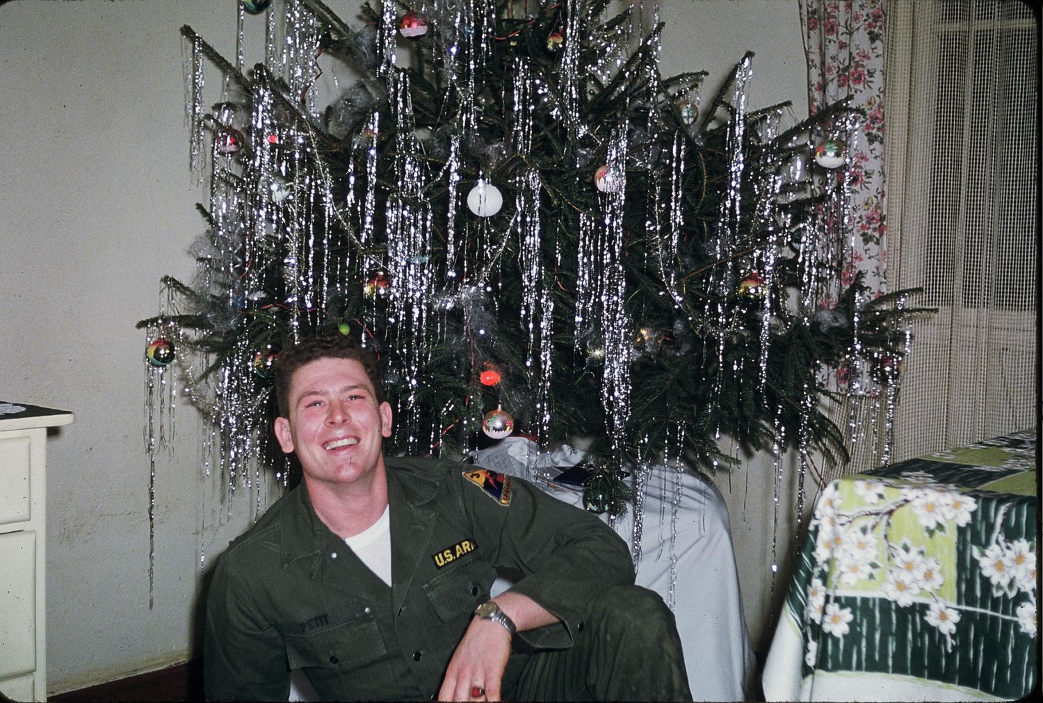 Smiling soldier in green military uniform sitting in front of a decorated Christmas tree with silver tinsel.