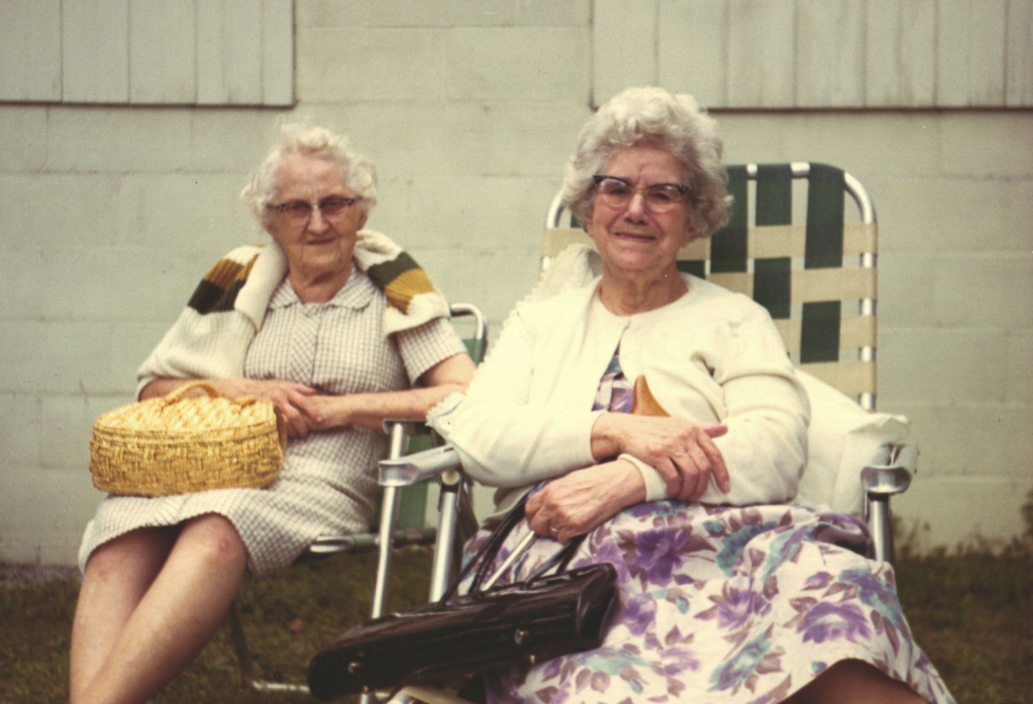 Two elderly women sitting outdoors in lawn chairs, one holding a wicker basket, smiling on a sunny day.