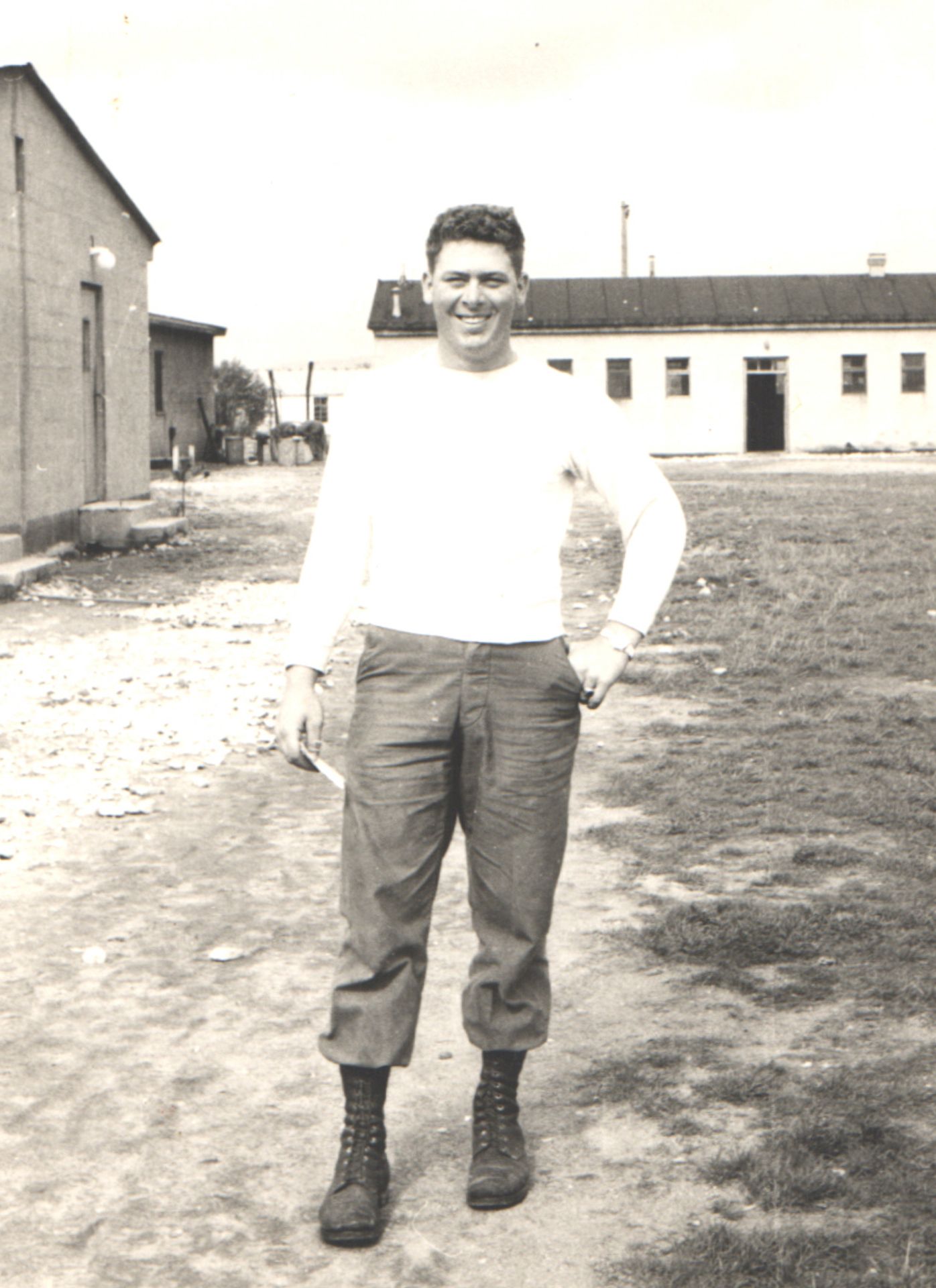Vintage black and white photo of a smiling man in military boots and casual clothes standing outdoors near barracks buildings.