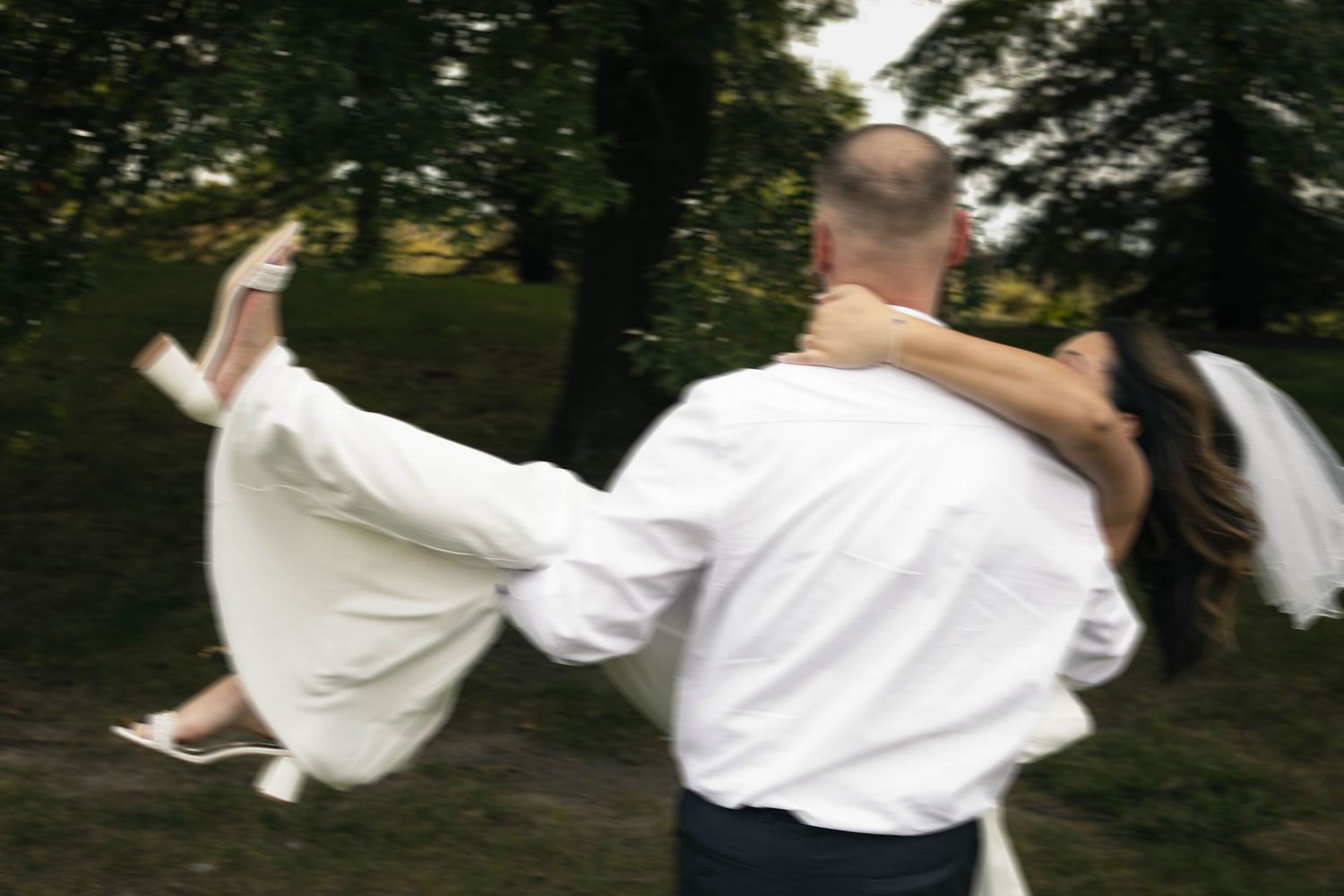 SAINT JAMES PHOTO- Groom in white shirt lifts bride in wedding dress outdoors in green park setting.