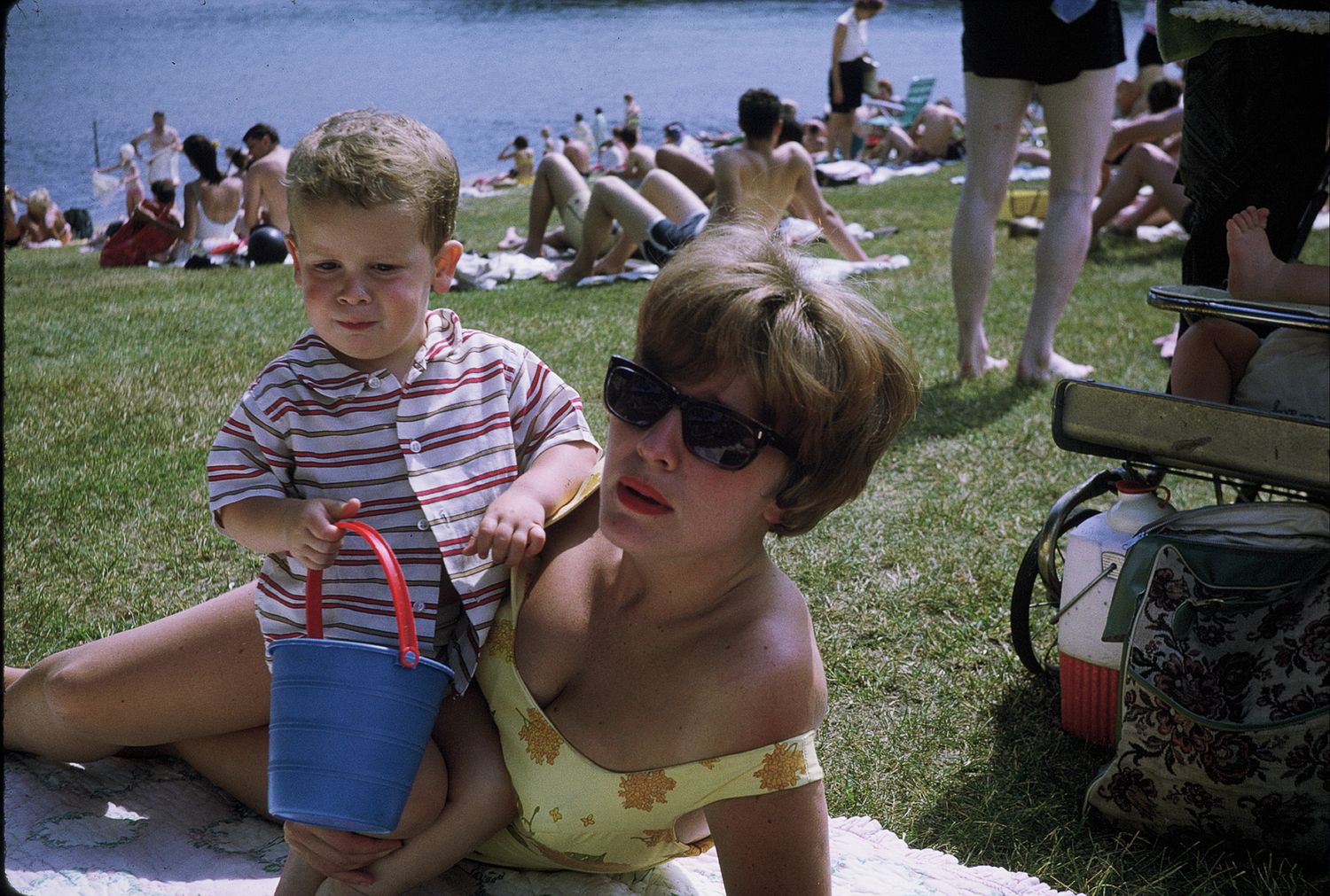 A young boy and a woman in sunglasses relax on grass near a lake at a sunny outdoor event.