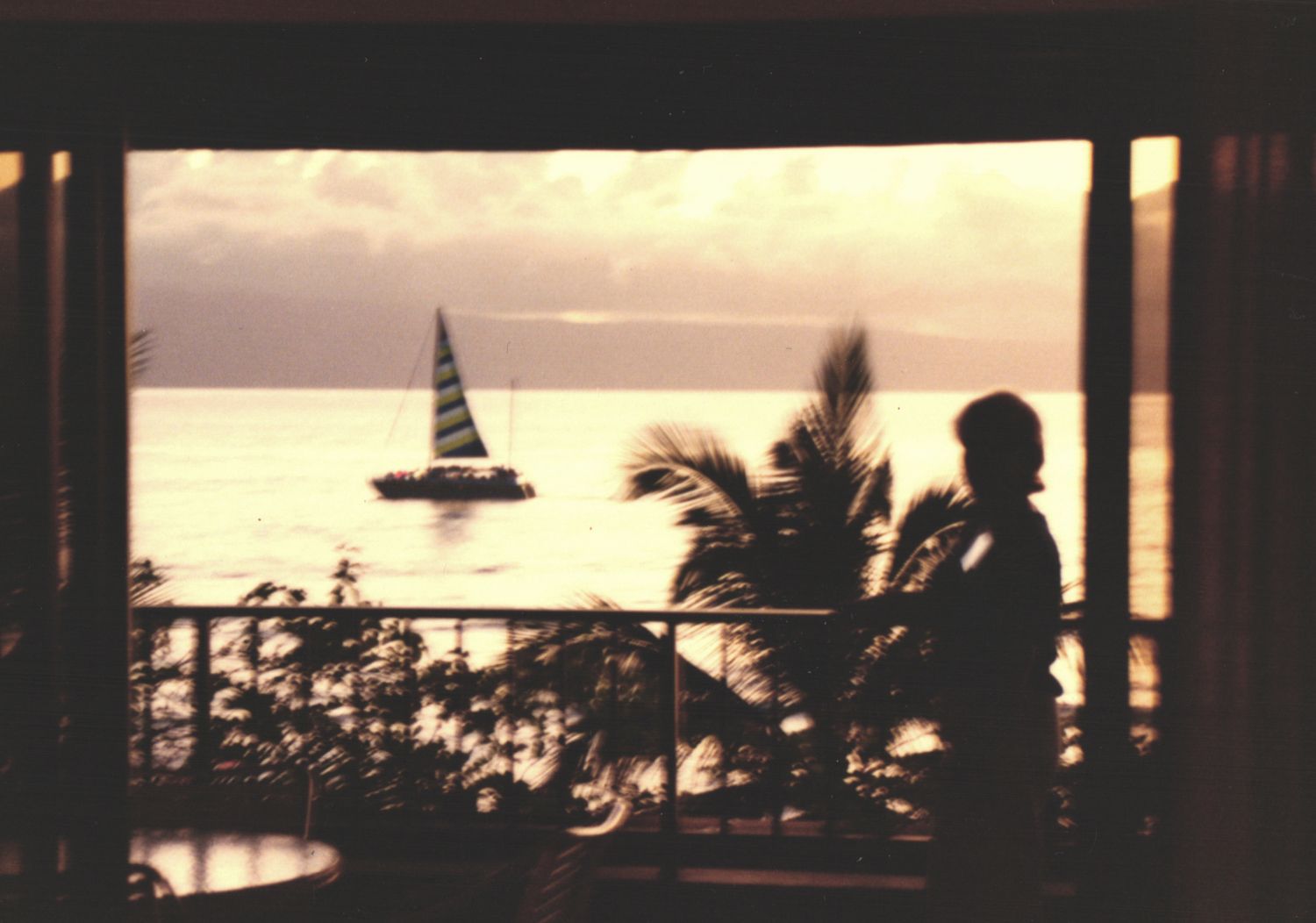 A child stands in silhouette on a balcony watching a sailboat at sunset over calm waters.