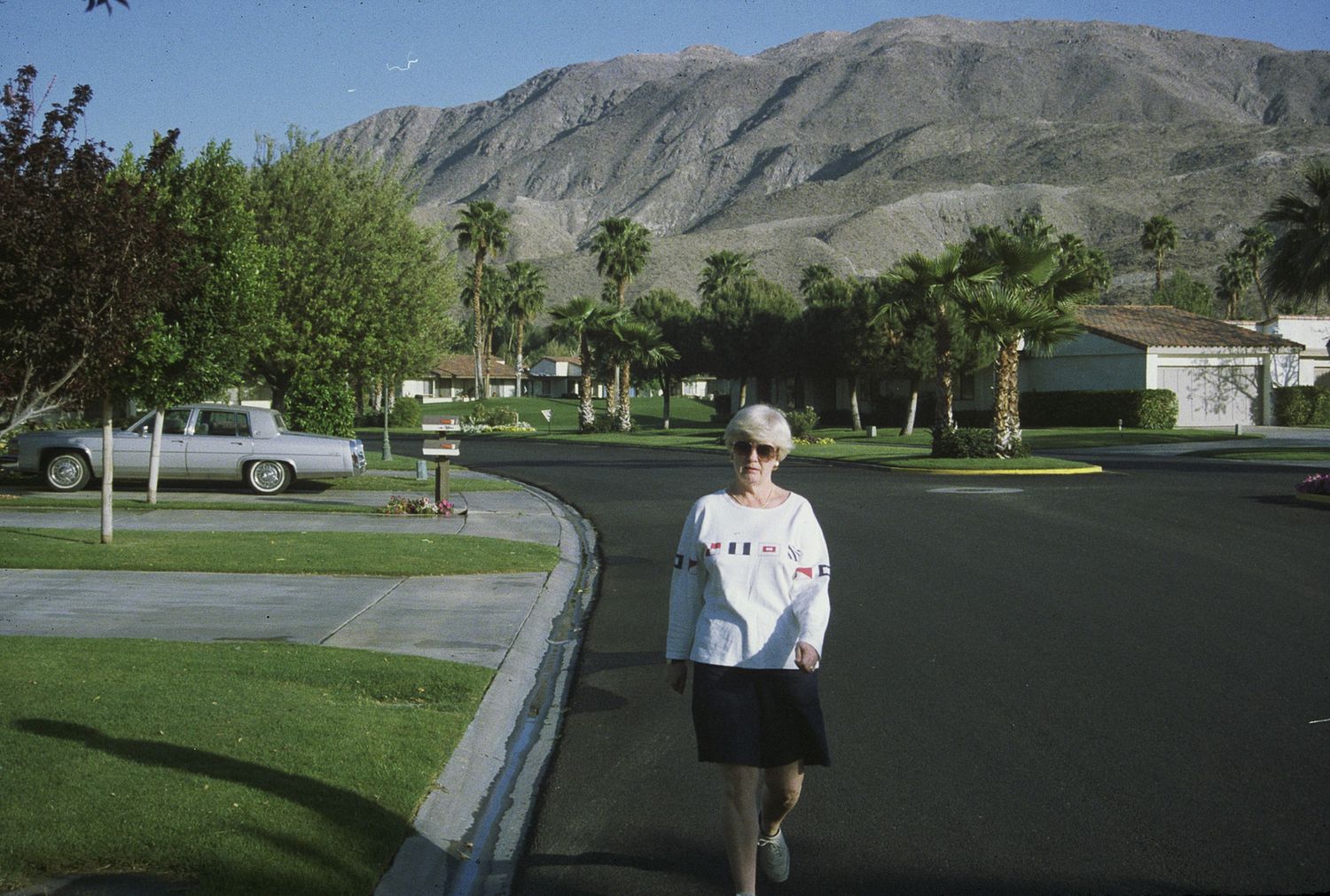 Person walking on a suburban road with palm trees and a mountain backdrop in a sunny desert community.