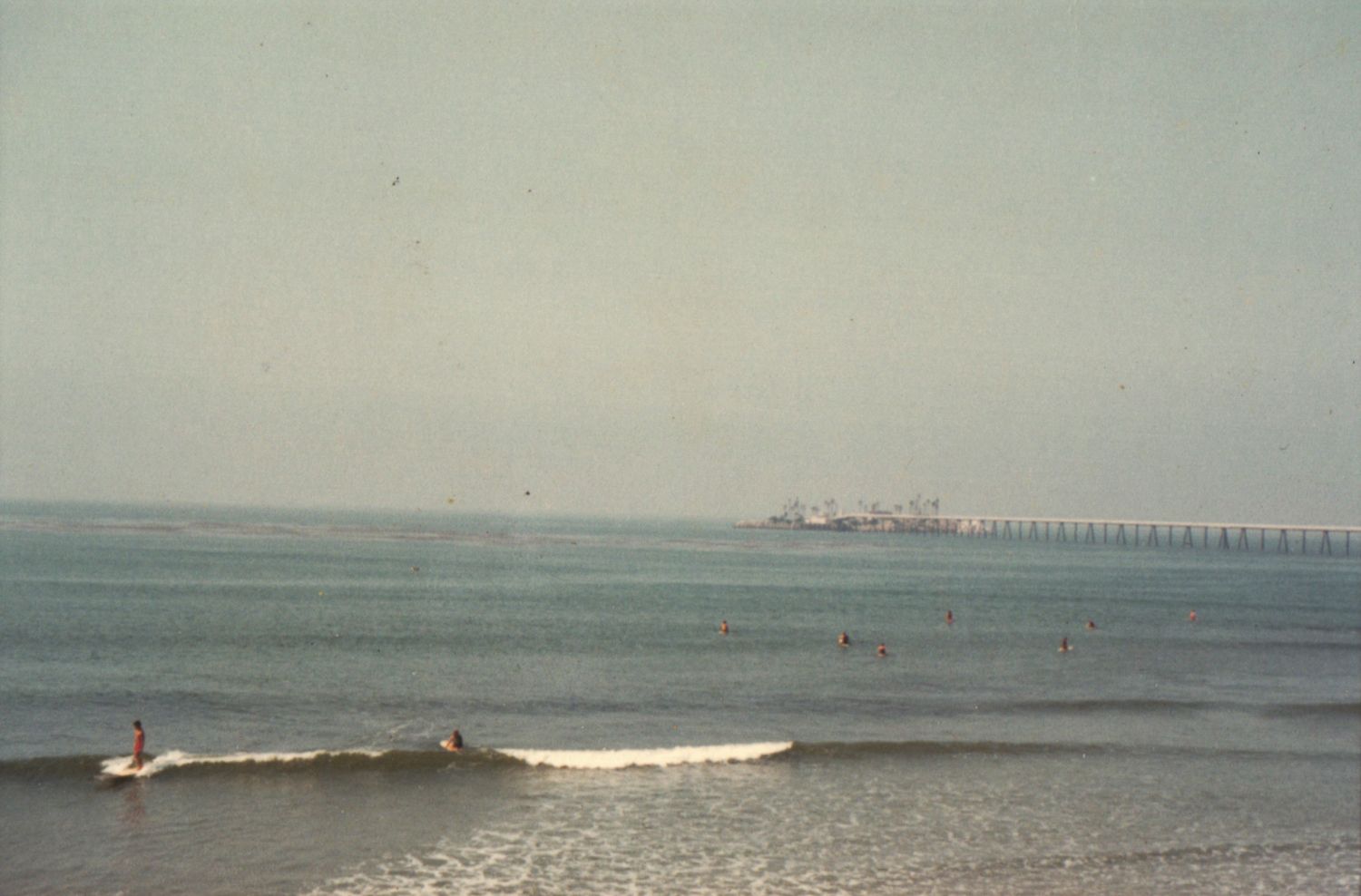 Surfers riding gentle ocean waves near a long pier stretching into the calm sea on a hazy day.