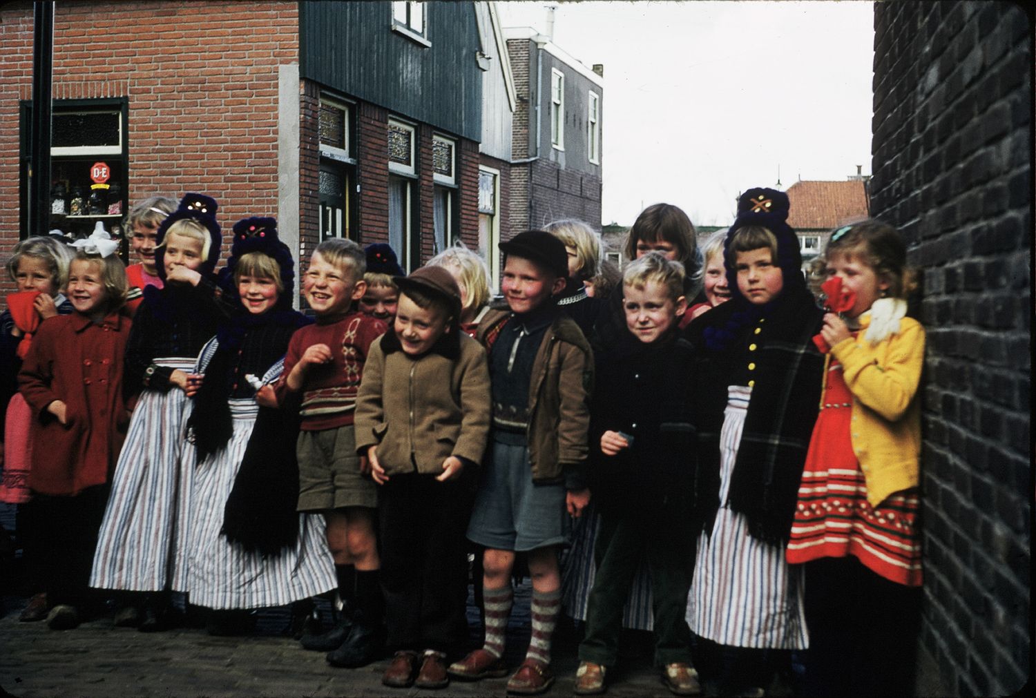 Group of Dutch children in traditional folk costumes lined up along a brick building street.