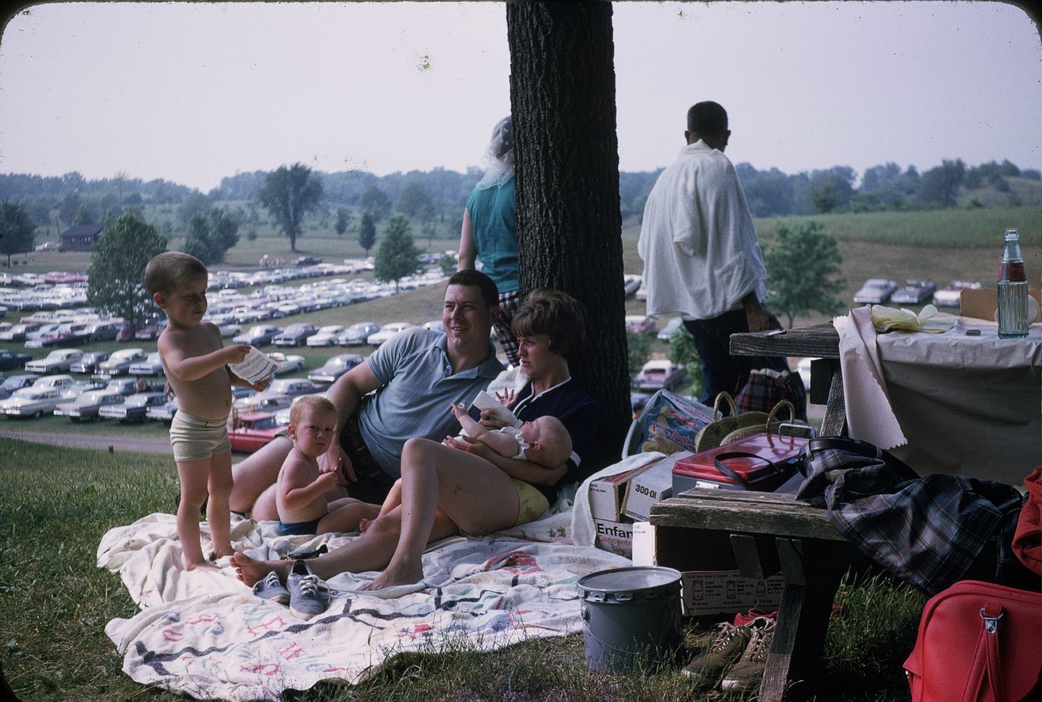 Family relaxing on blanket under tree at outdoor event with large parking lot in background.