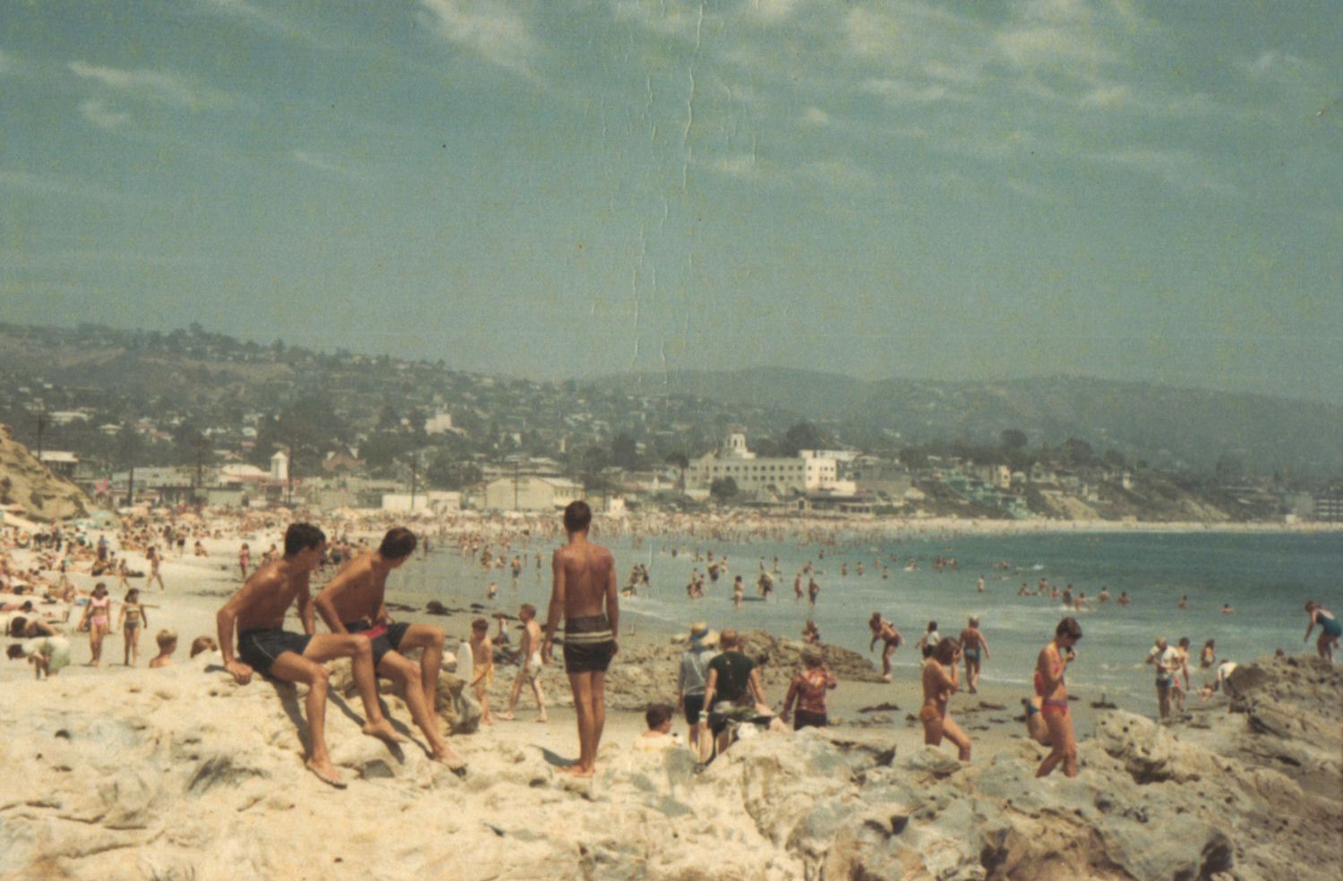 Crowded beach scene with swimmers and sunbathers on rocky shore with ocean and hillside town in background.