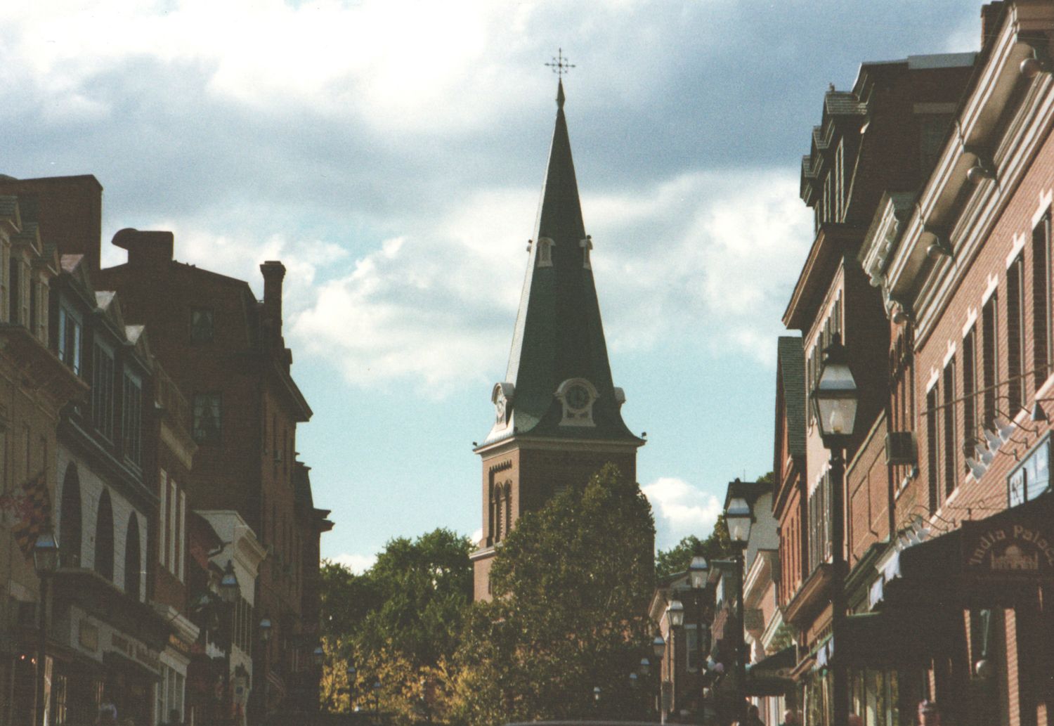 Historic town street with brick buildings leading to a classic church steeple under a dramatic cloudy sky.