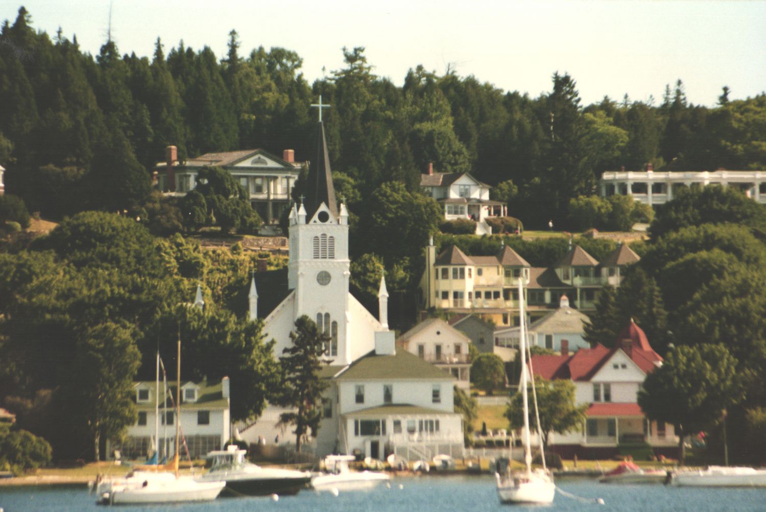 A picturesque waterfront town with a white church steeple, colorful buildings, and sailboats on calm blue water.