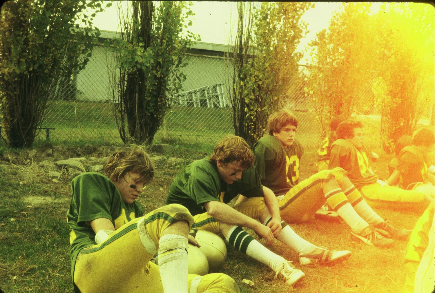 Football players in green and yellow uniforms rest on the ground during a game break outdoors.