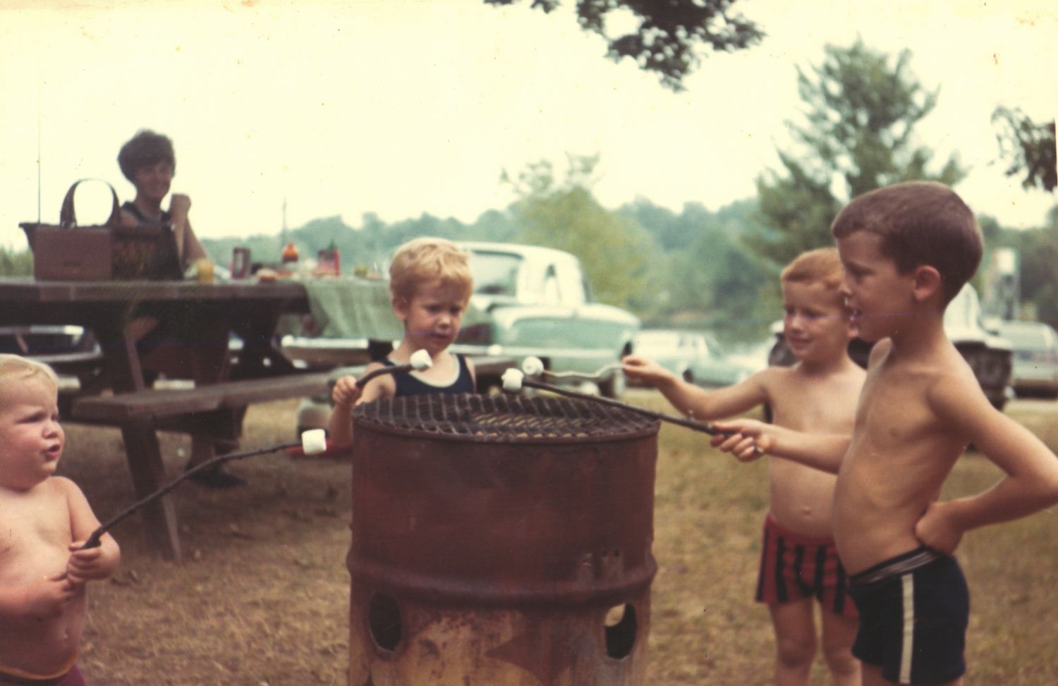 Children roasting marshmallows over a barrel grill at a campground in a vintage 1960s outdoor family gathering.