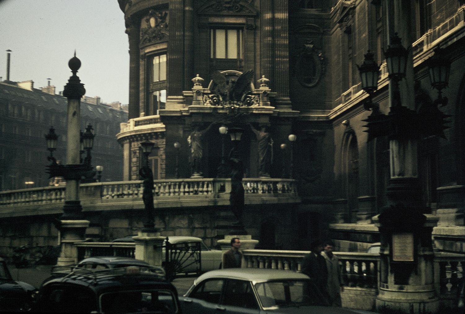 Historic European city street with ornate classical architecture, balustrades, and vintage cars below.