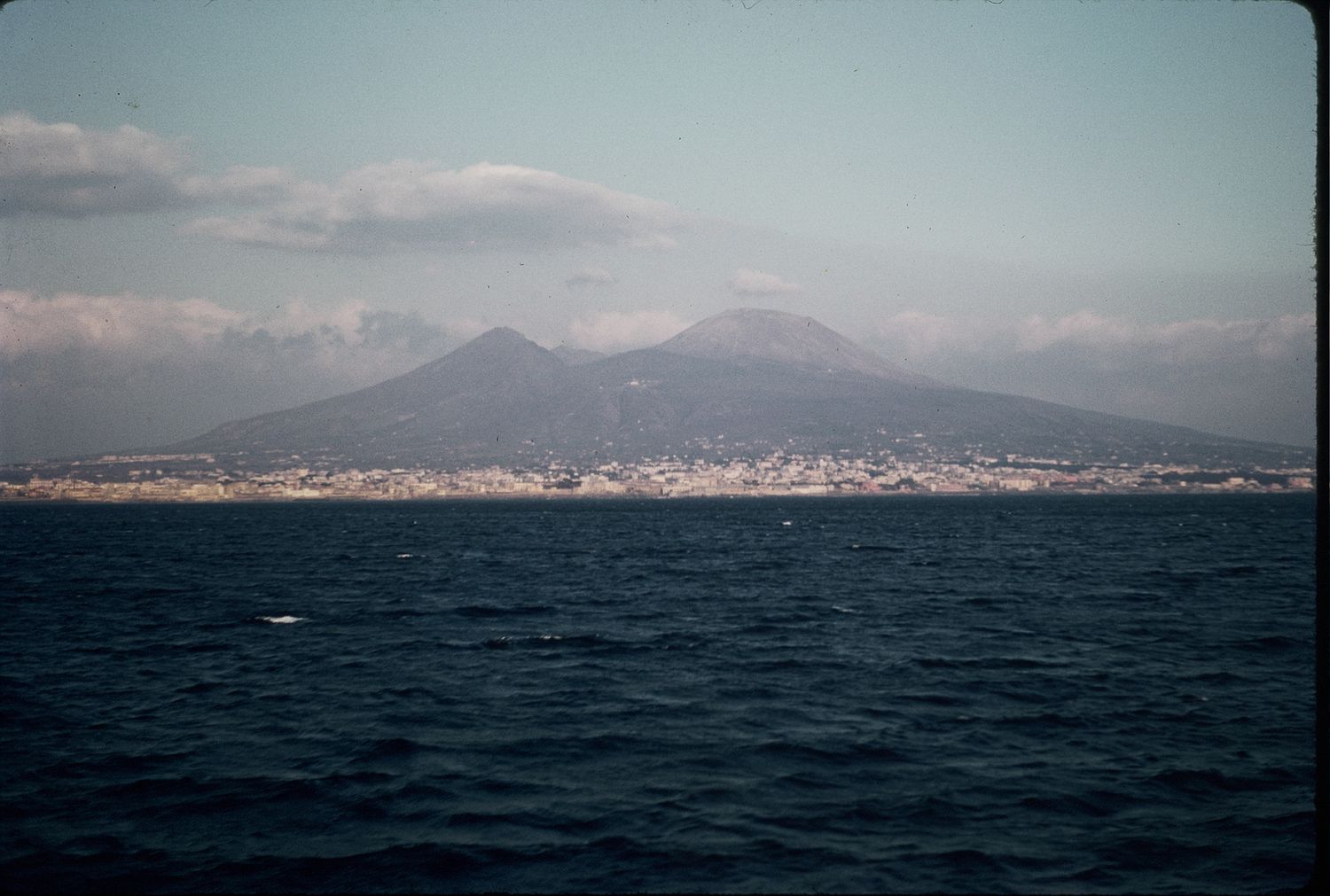Distant view of Mount Vesuvius rising above Naples cityscape across the calm blue Mediterranean Sea.
