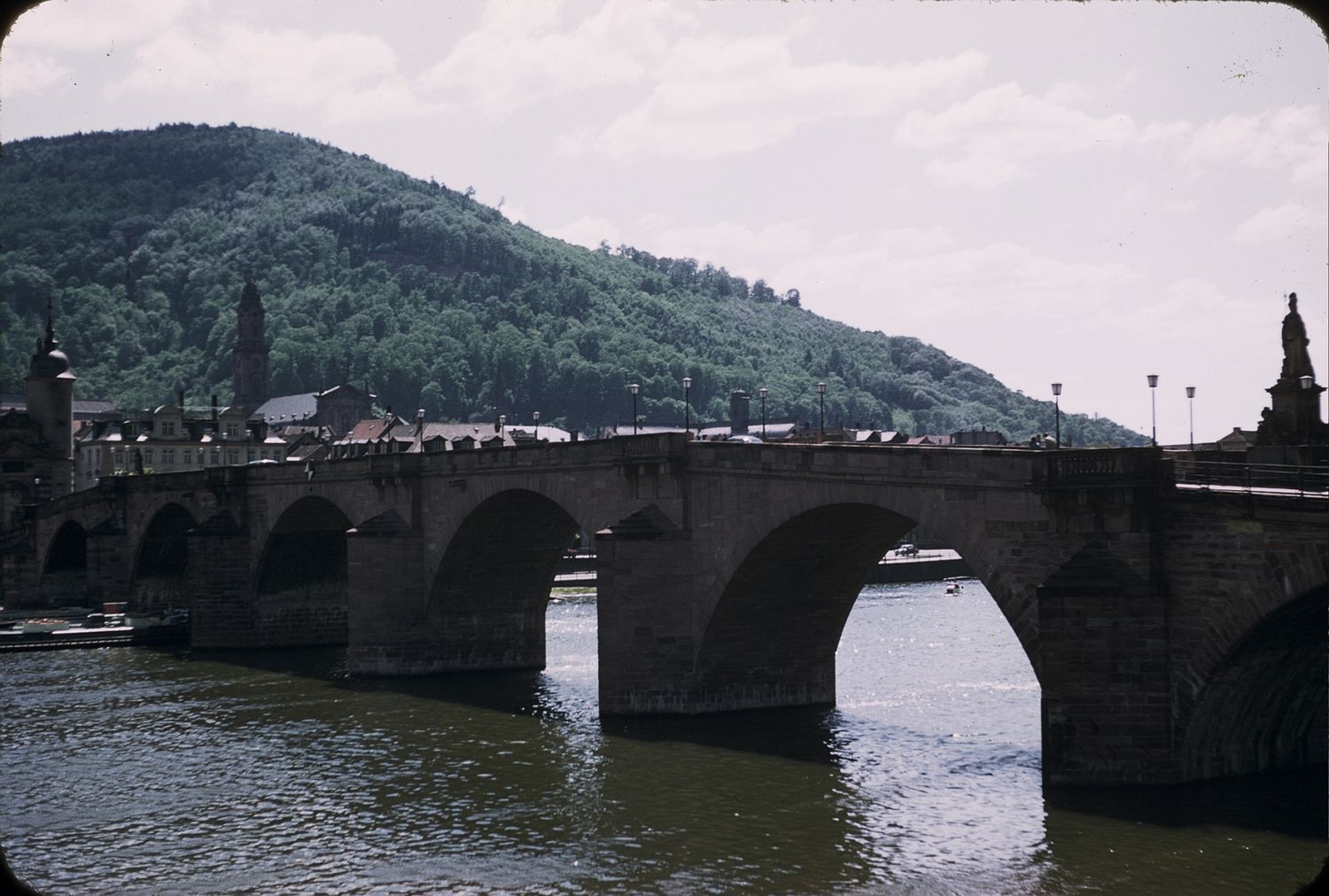 Historic stone arch bridge spanning a river in Heidelberg, Germany, with a forested hill and city skyline in the background.