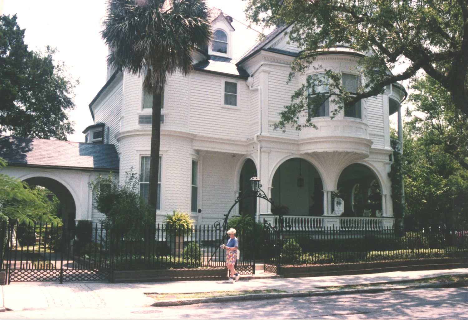 A grand white Victorian mansion with arched portico and iron fence, surrounded by lush trees in a historic neighborhood.