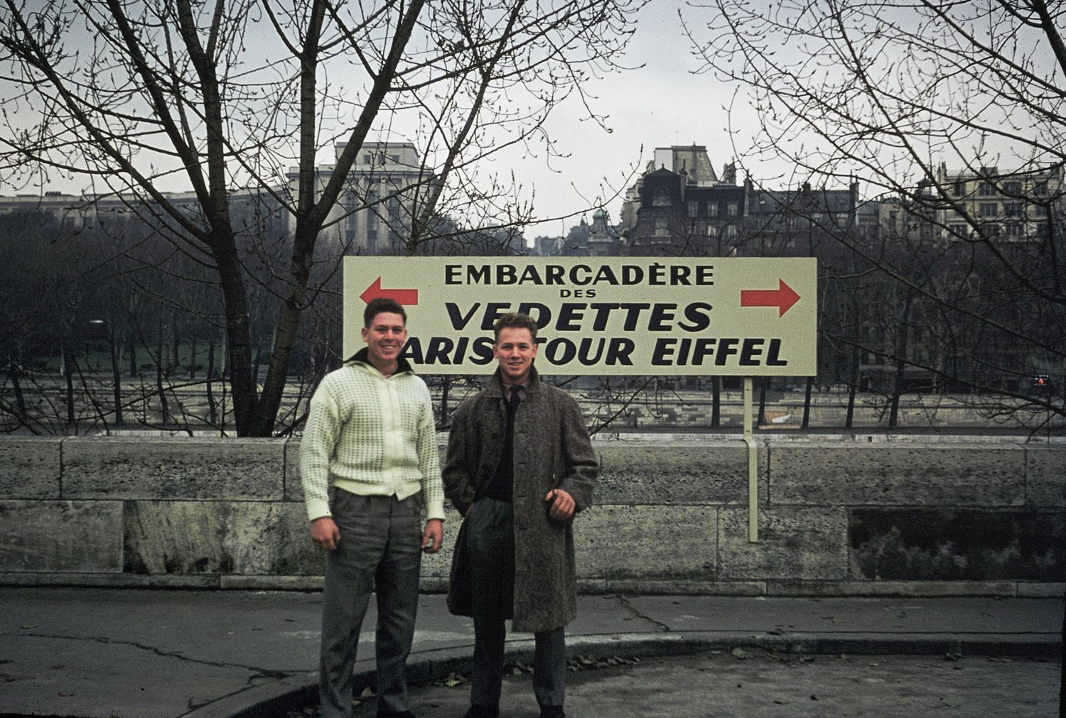 Two men posing in front of Vedettes Paris Tour Eiffel embarcadere sign along the Seine River.