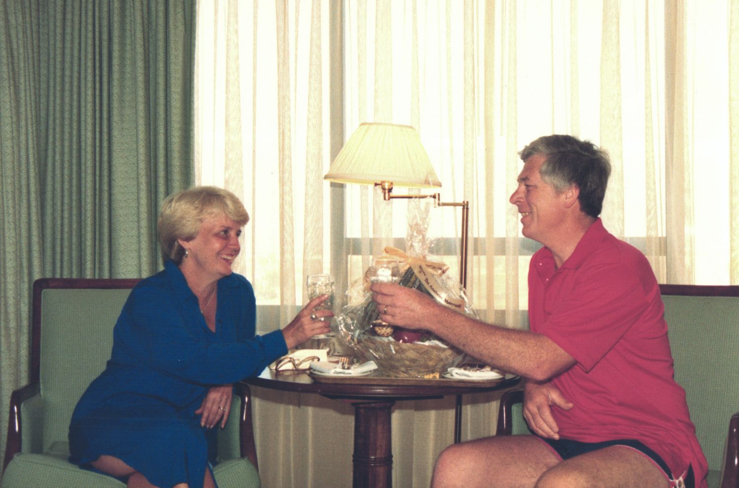 A man and woman toasting drinks at a small table in a warmly lit hotel room.