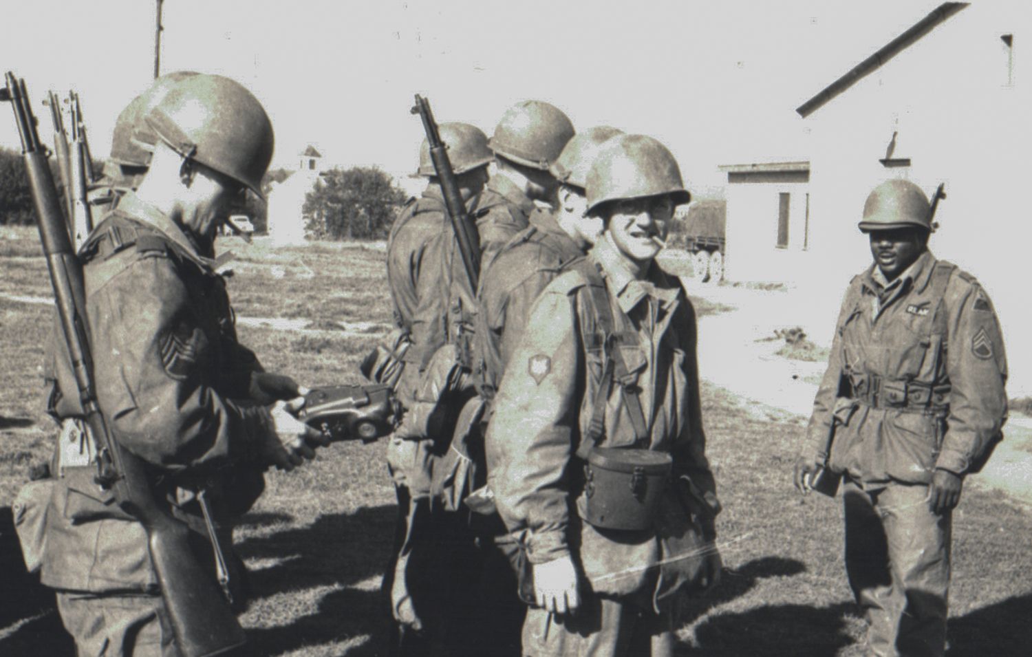 Black and white photo of soldiers in helmets and gear standing outside military barracks.