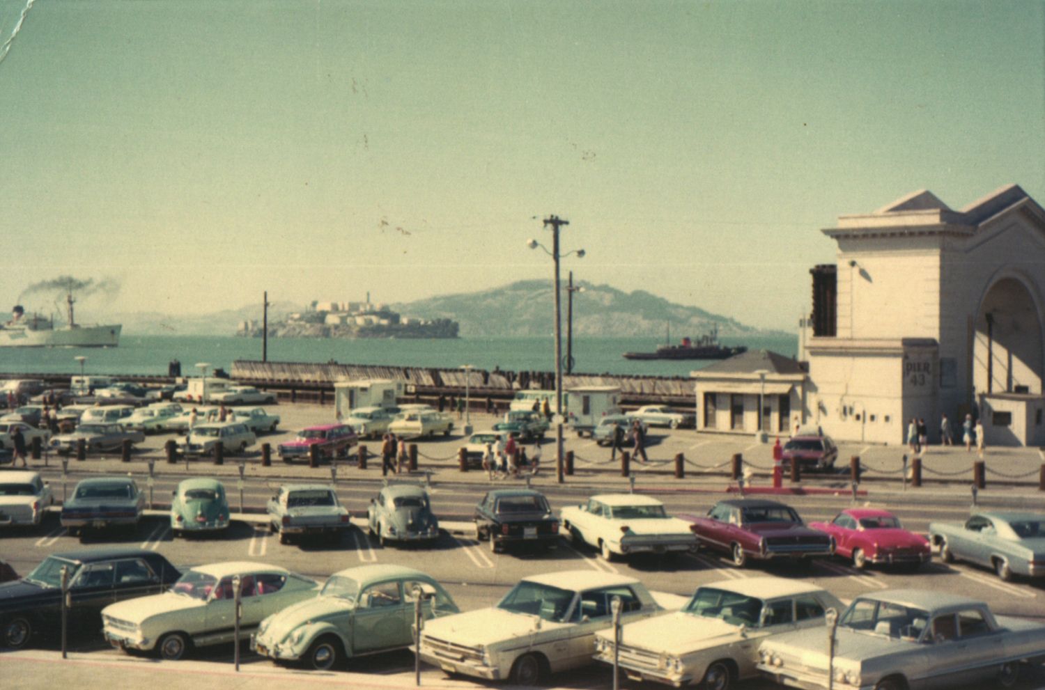Vintage 1960s waterfront parking lot with classic cars near a harbor, ships, and mountains in the background.