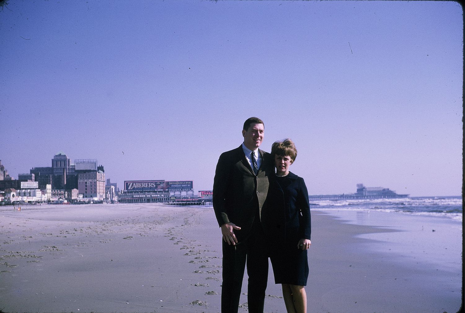 A couple stands together on a sandy beach with a coastal city skyline and ocean waves visible in the background.
