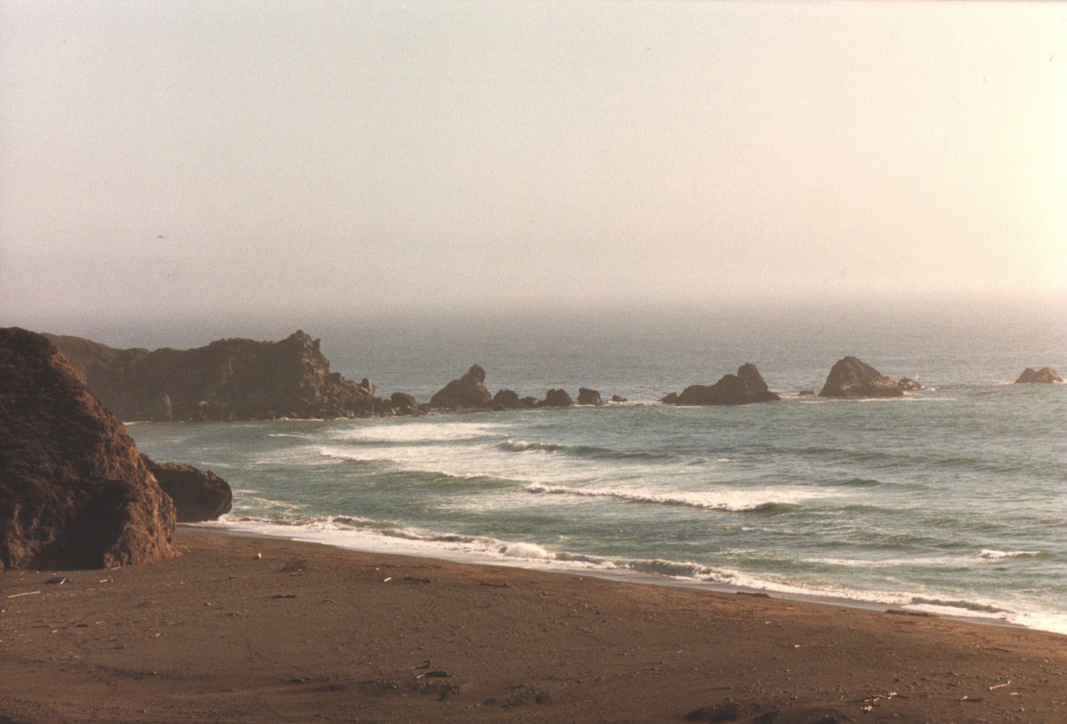 Misty coastal beach scene with rocky sea stacks emerging from the ocean waves along a sandy shore.