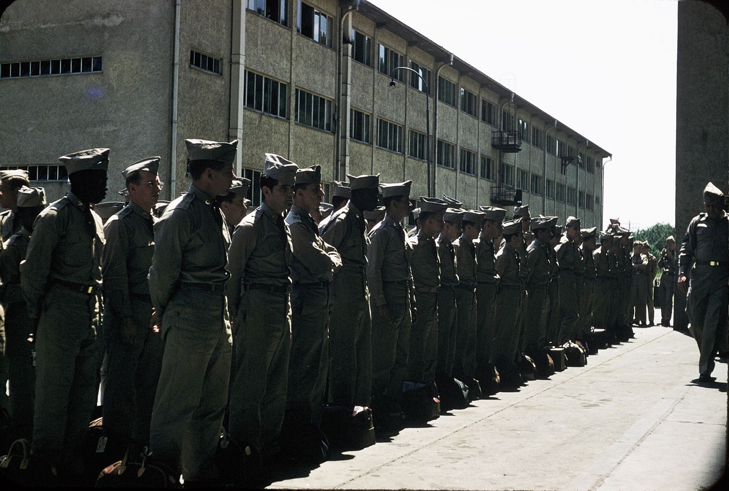 Rows of soldiers in uniform standing in formation outside a large military building on a sunny day.