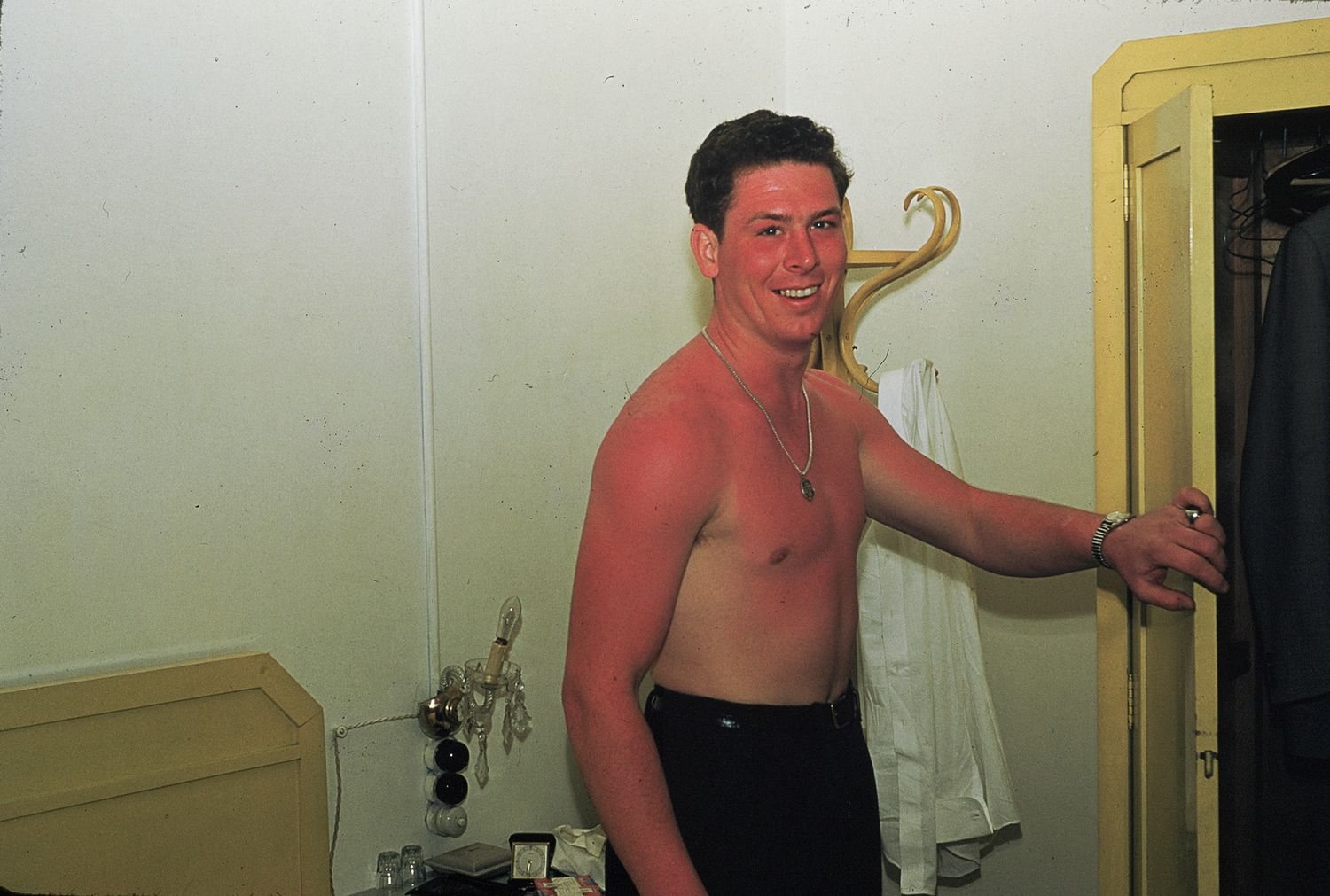 Shirtless smiling man standing in a room, holding a door frame, wearing dark pants.