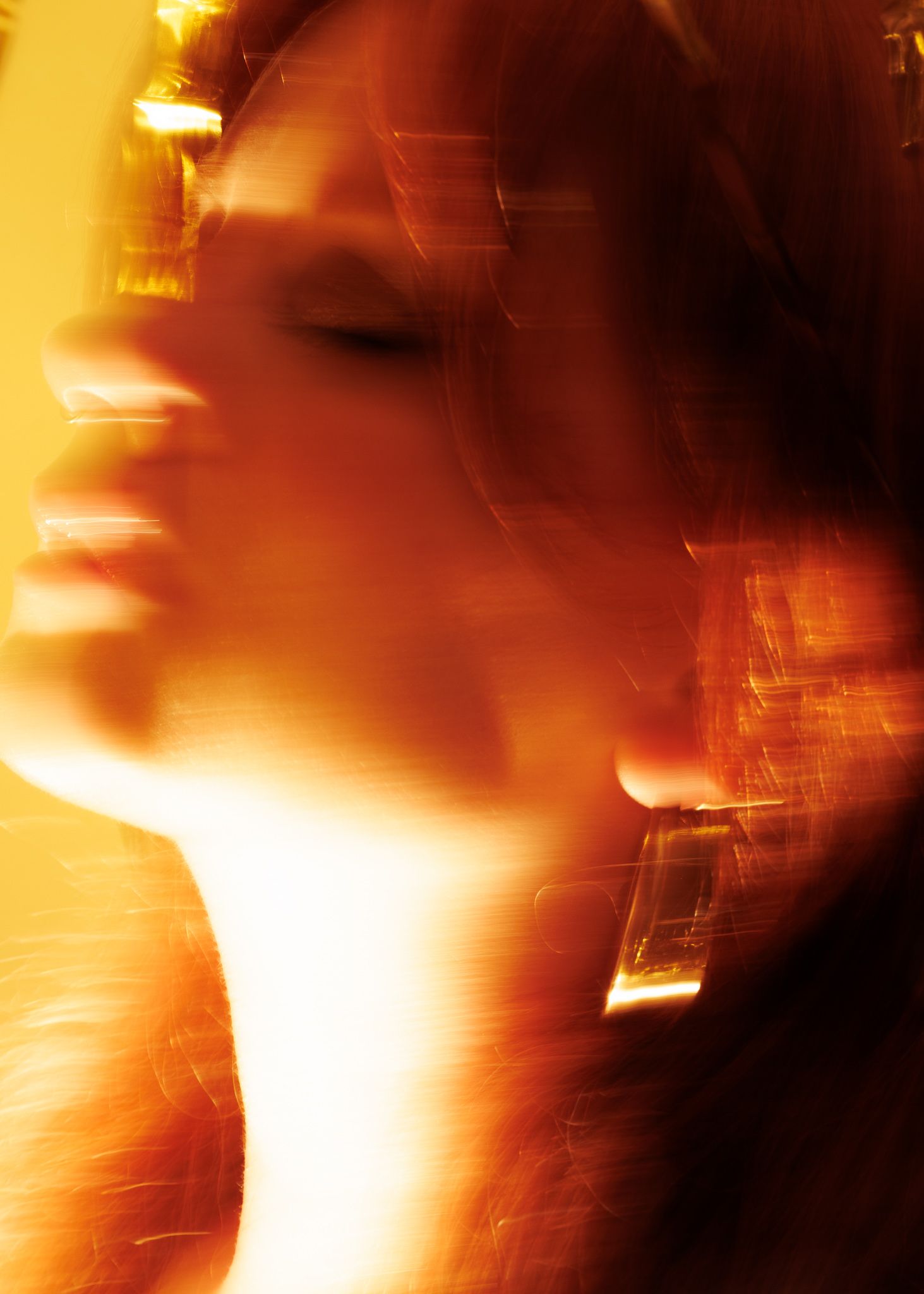 A blurred artistic portrait of a woman bathed in warm golden and orange light with eyes closed.