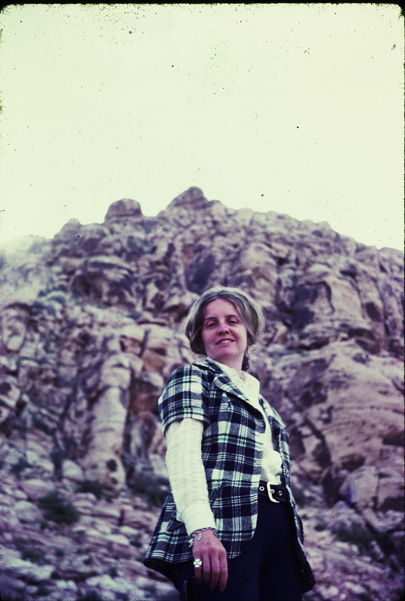 Woman in plaid vest smiling before rocky mountain landscape backdrop outdoors.