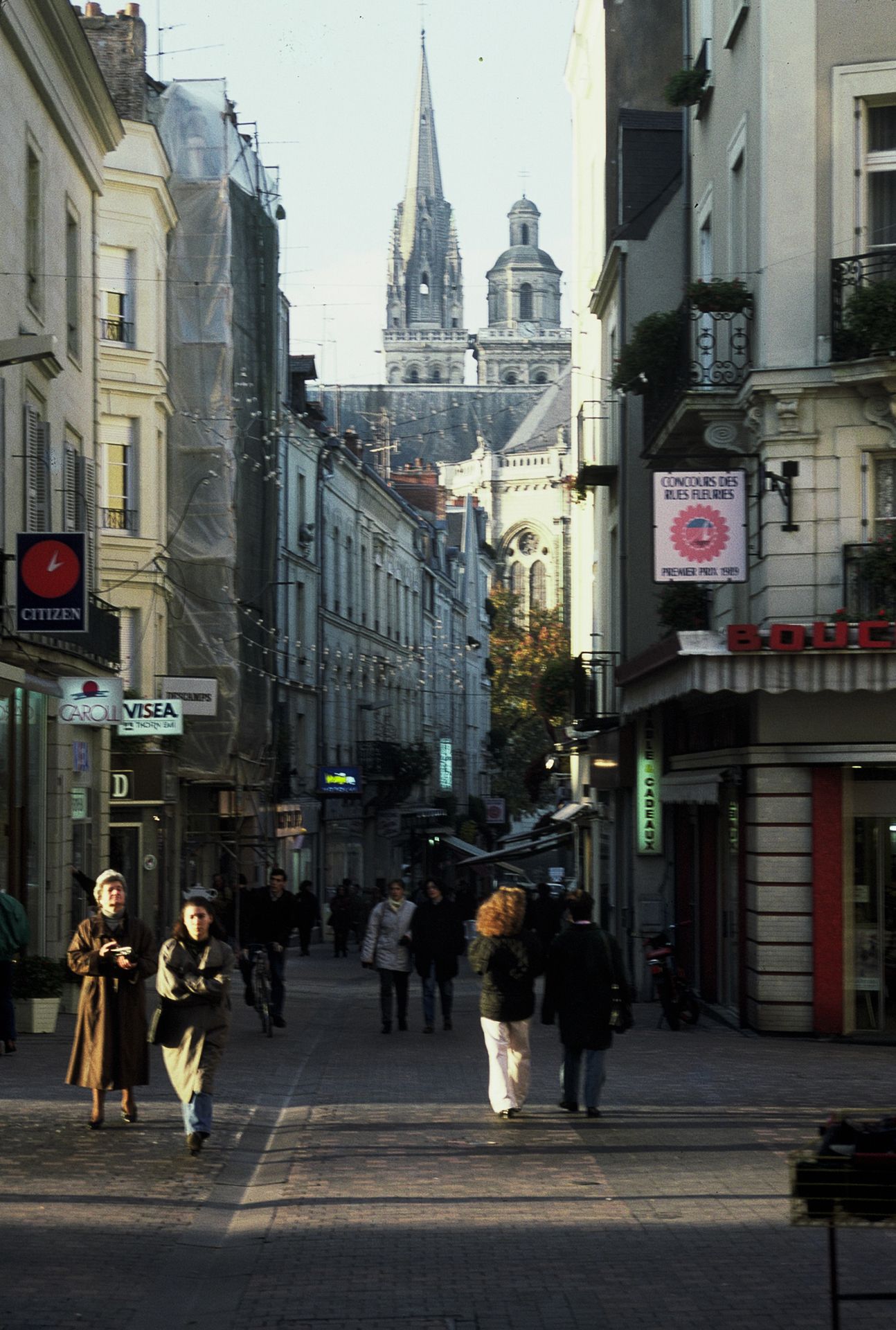 Busy pedestrian shopping street in Angers, France, with Gothic cathedral spires visible in the background.
