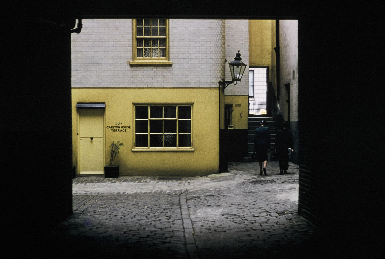 Cobblestone alley framed by a dark doorway leading to a yellow building with vintage windows and a gas lamp.
