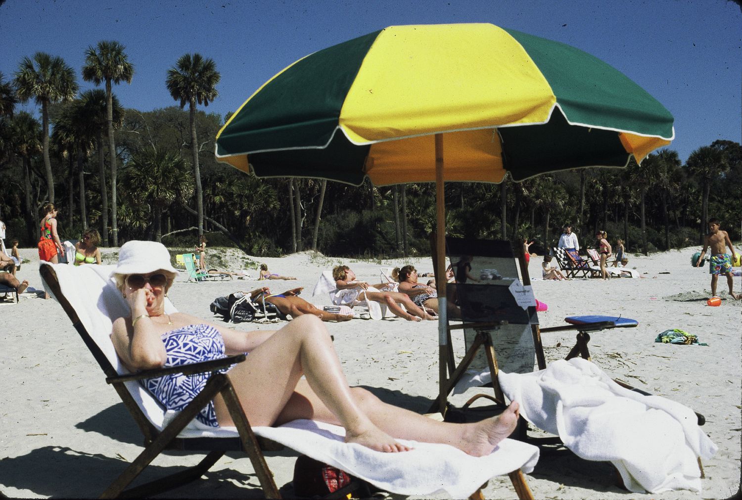 Woman relaxing in lounge chair under colorful beach umbrella on sunny sandy beach with palm trees.