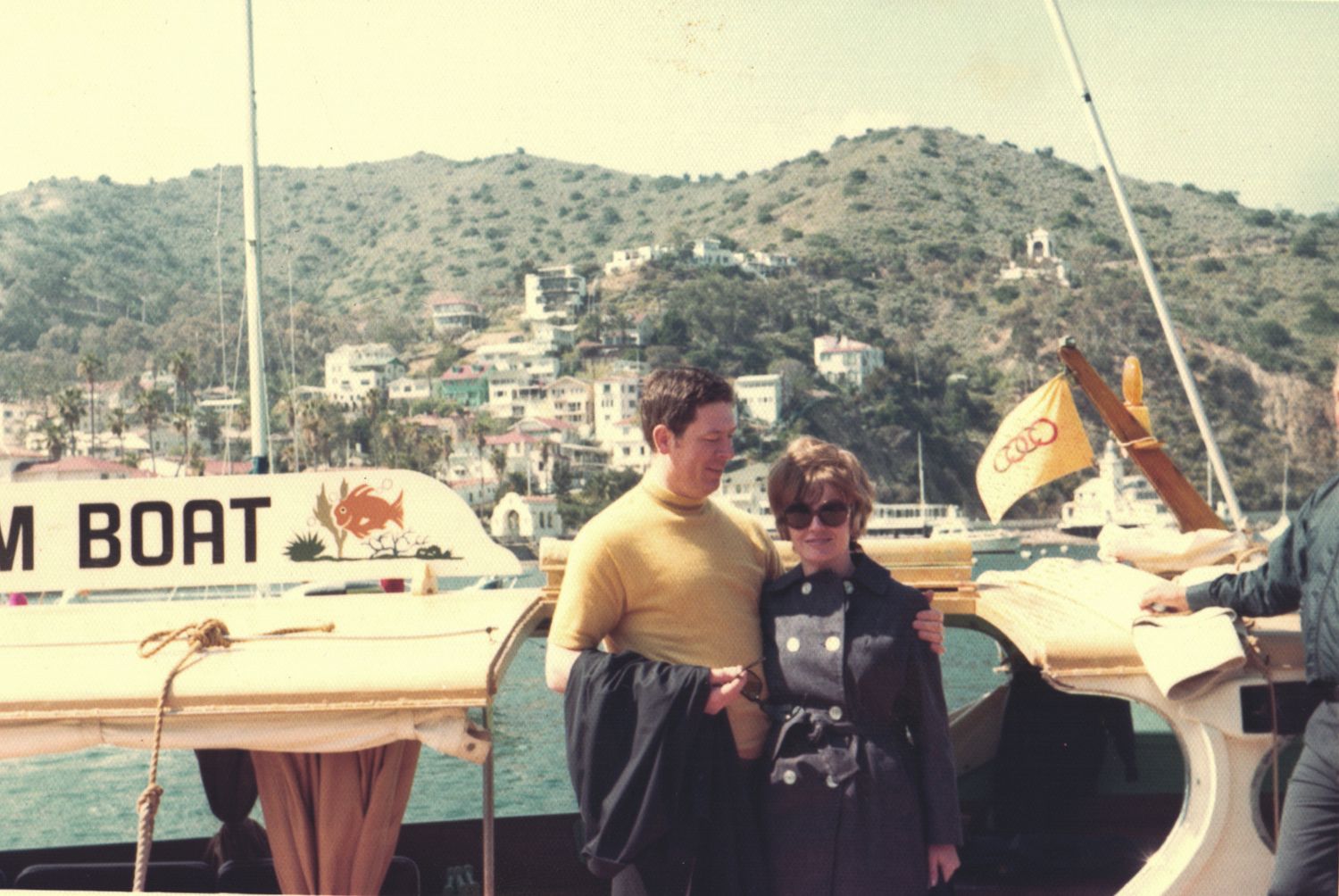 Couple posing on a boat dock with sailboats and hillside town in background, vintage 1960s photo.
