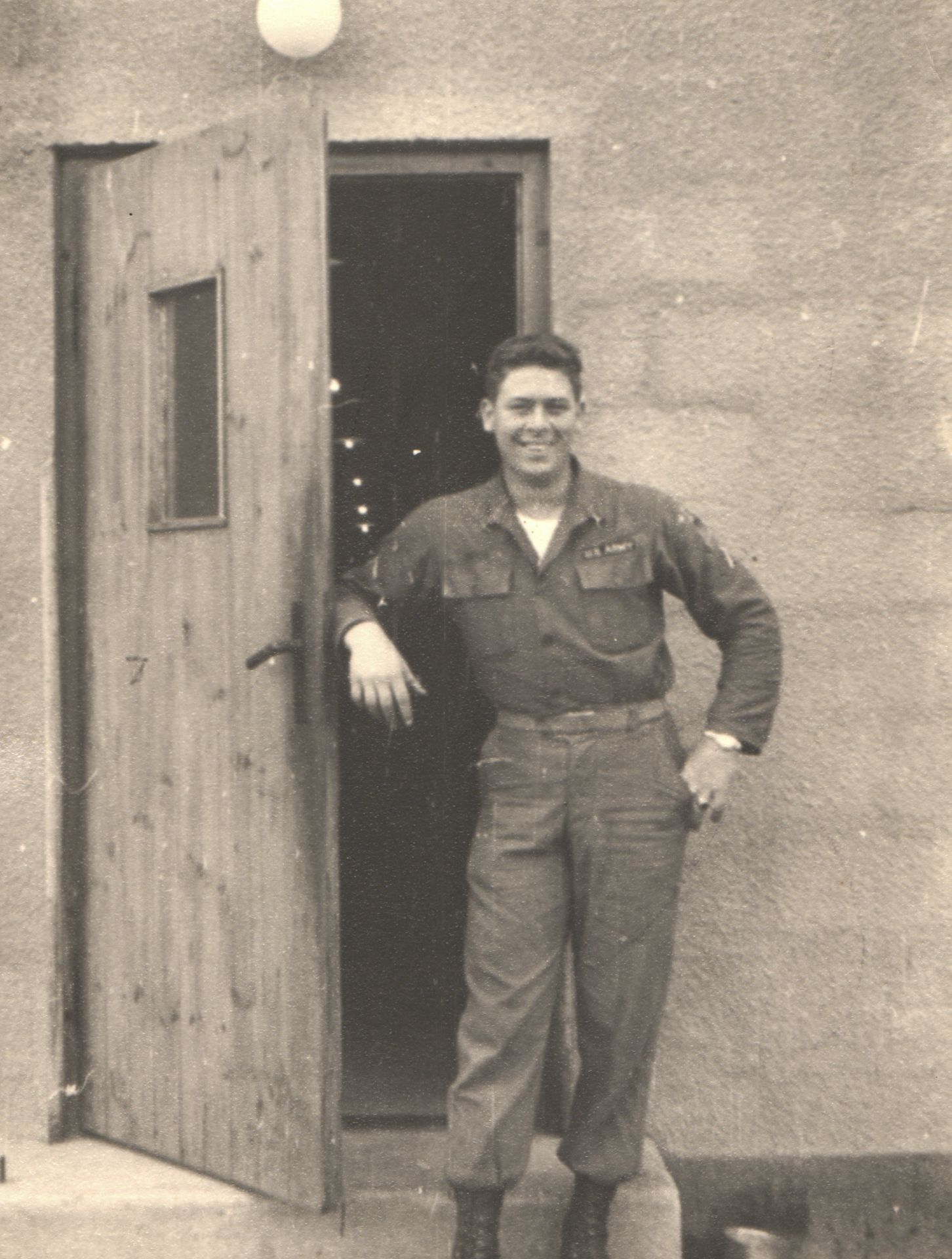Vintage black and white photo of a smiling U.S. Army soldier in uniform standing by a wooden door.